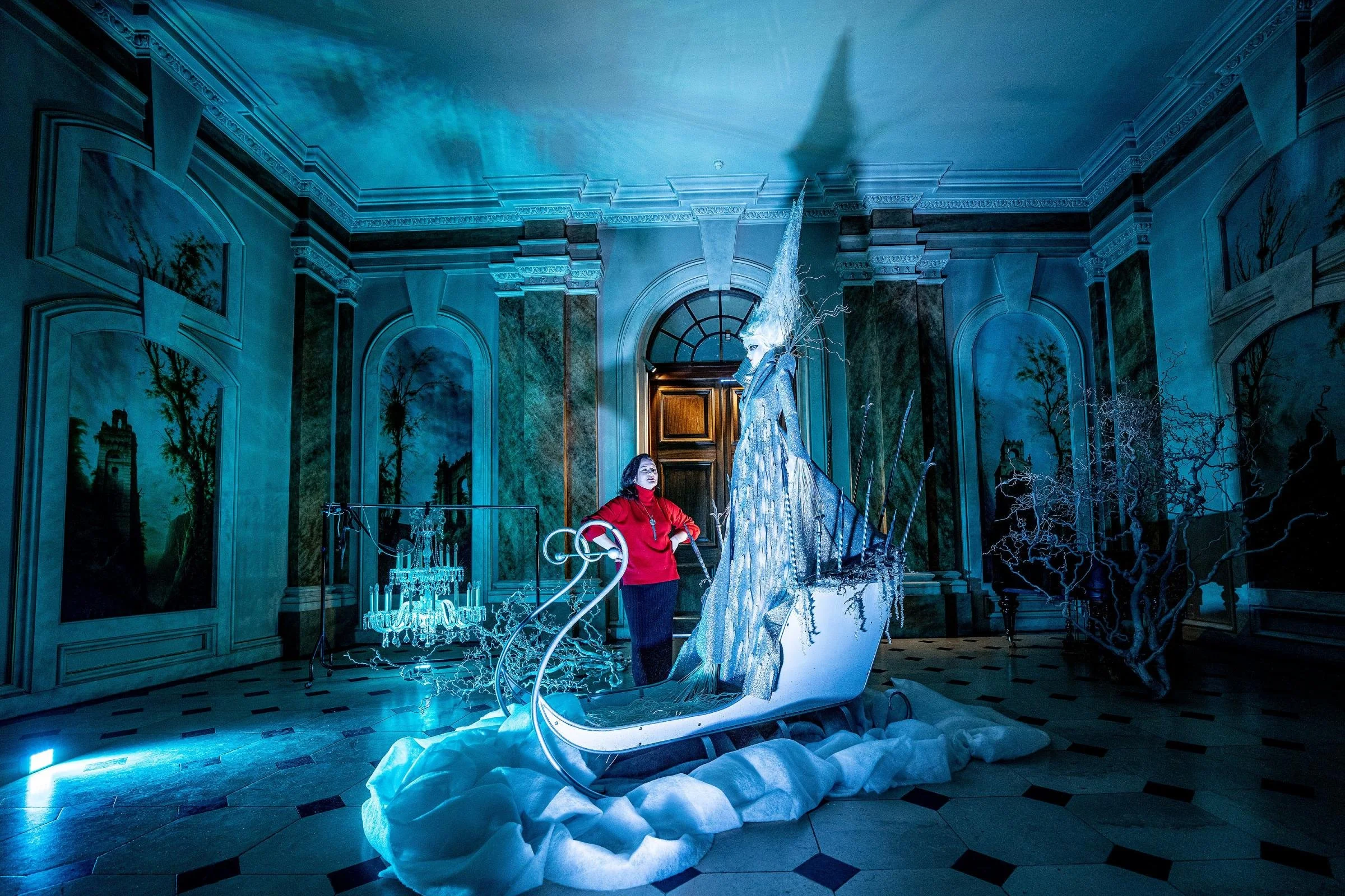 An ornate room with a high ceiling, detailed moldings, and painted wall murals of trees and arches. A woman in a red top and black pants stands near an elaborate, illuminated sculpture that resembles a mythical creature, with lightning bolt-like elem
