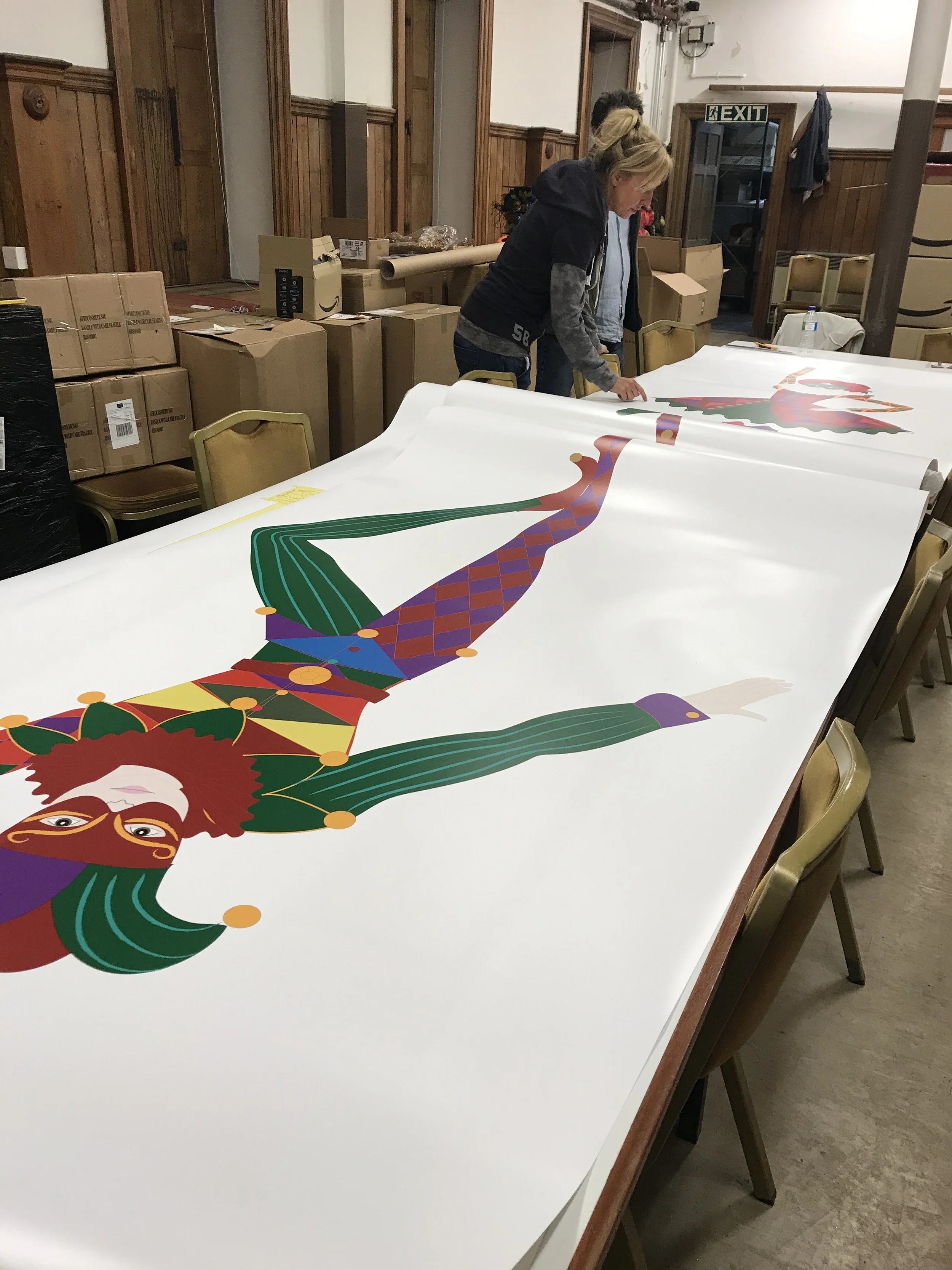 Two women examining a large, colorful, cartoon-style illustration of a jester or clown spread out on a table in a room with wood-paneled walls and boxes stacked in the background.