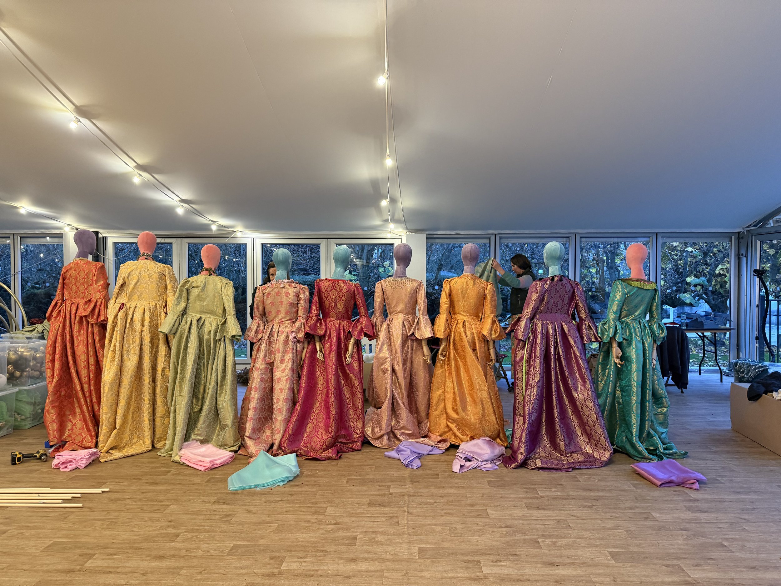 A row of eleven mannequins dressed in colorful, intricate gowns displayed on a wooden floor with fabric pieces scattered in front of them and a large window in the background showing trees outside. There are two women adjusting the mannequins' outfit