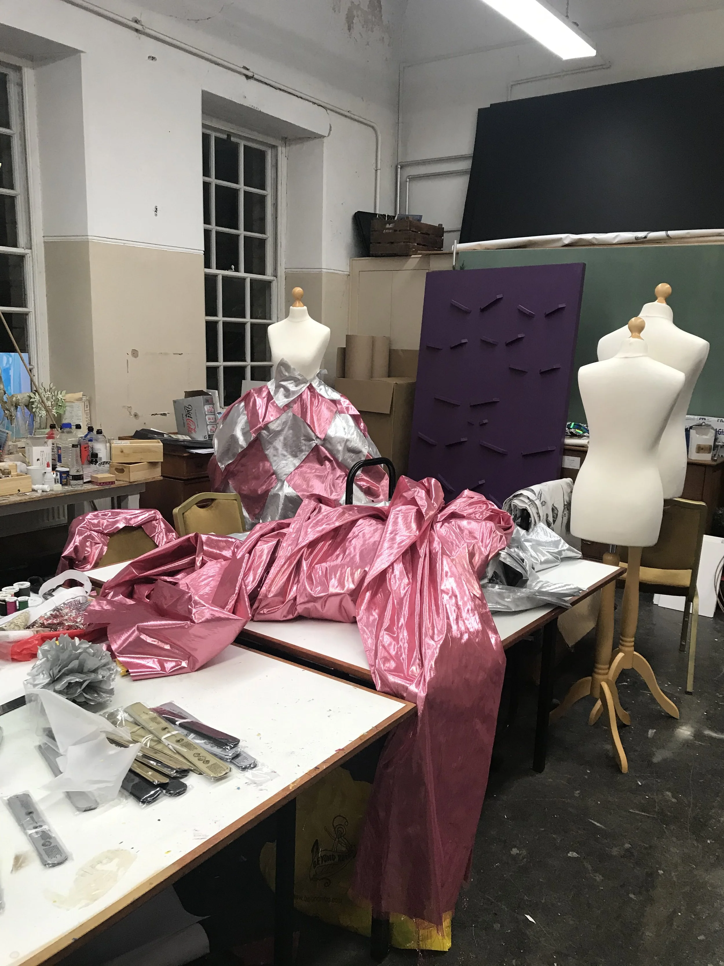 A room with mannequins dressed in pink and silver fabric, likely in a workspace or studio, with tables covered in sewing supplies, fabric, and papers.