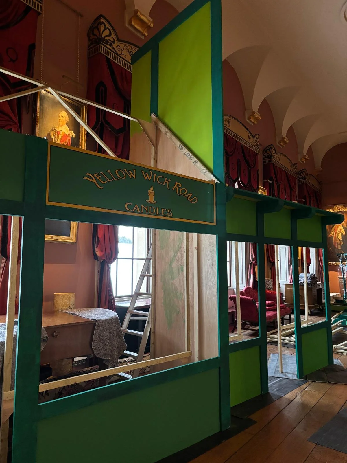 The image shows a partially constructed set for a production, resembling a scene from a storybook or themed environment. It features a green frame with a sign that reads 'Yellow Wick Road Candles,' and depicts a room with pink walls, tall arched wind
