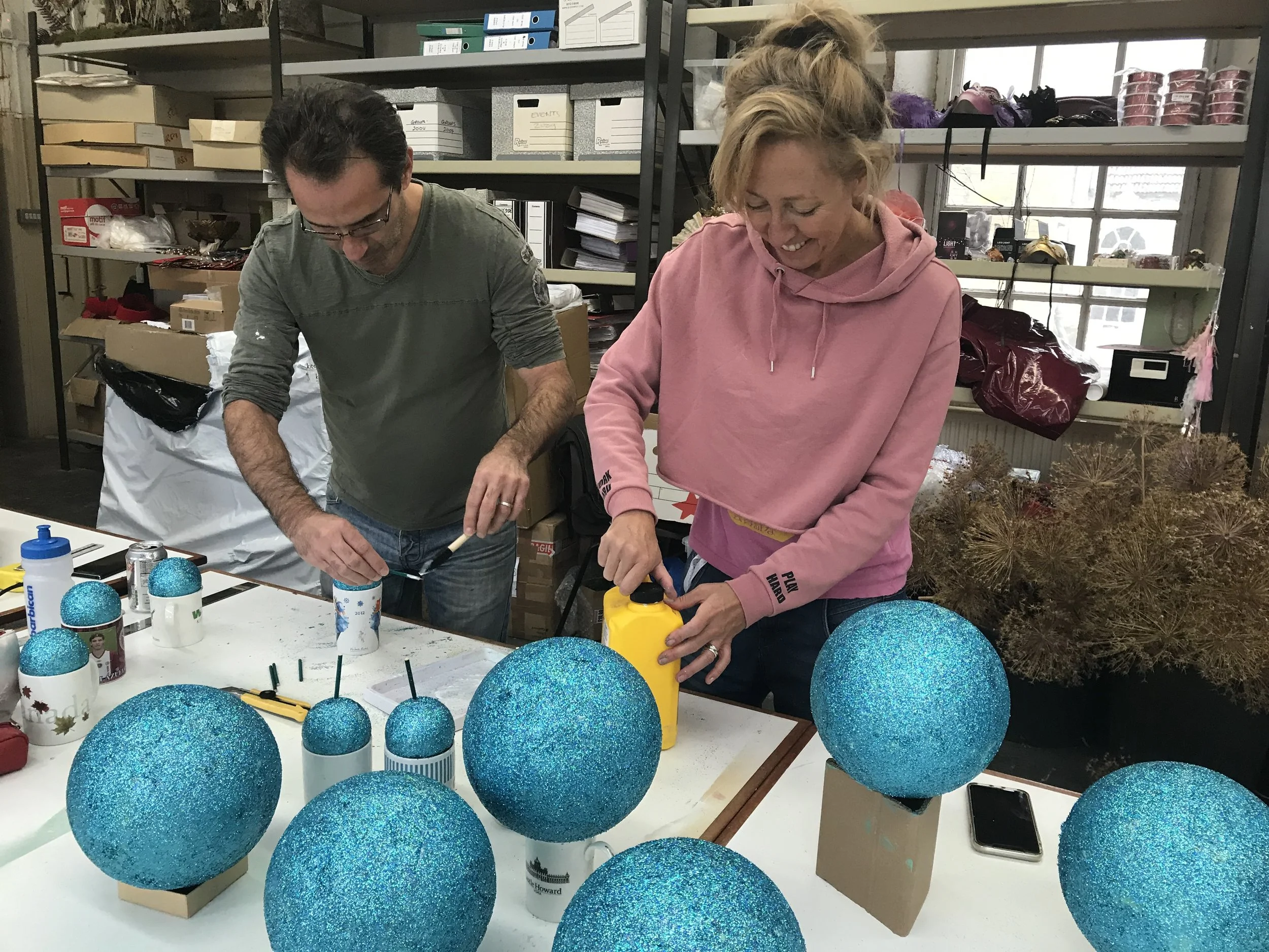 Two women are making holiday decorations in a craft room filled with shelves of supplies. They are surrounded by large blue glittery ornaments on a table, some with photo stickers, all placed in cups. The women are smiling and focused on their work.