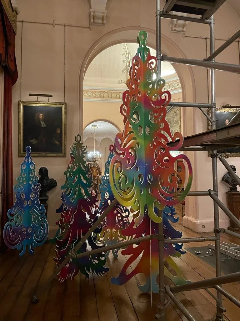 Colorful, ornate, cut-out decorative trees on display inside a museum with wooden floors, framed paintings, and scaffolding.