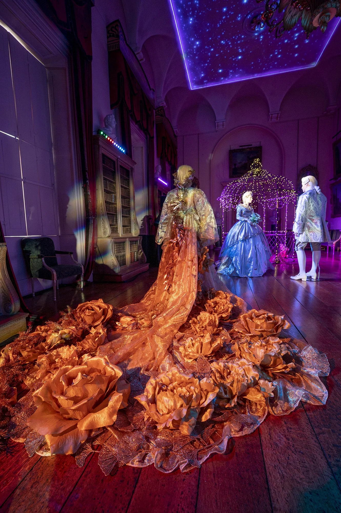 An indoor scene at a museum or gallery with mannequins dressed in elaborate, colorful costumes. The costumes are in vibrant orange and shiny blue. There is a ceiling with starry sky and purple lighting, along with decorative lighting and furniture.