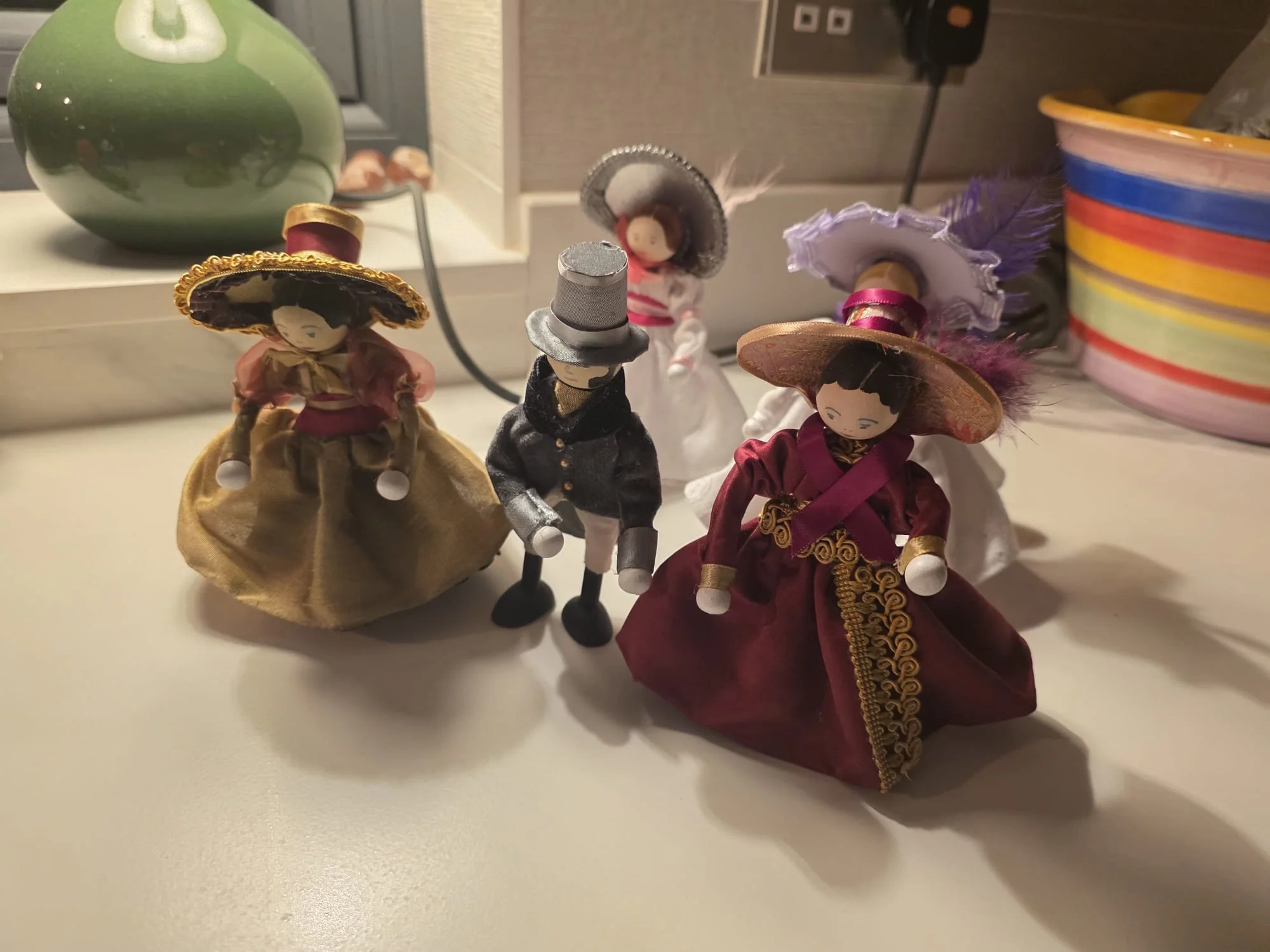 Four small decorative dolls dressed in traditional Mexican attire, with three women in colorful dresses and wide-brimmed hats, and one man in a gray suit with a top hat, standing on a white surface in front of a countertop with a green bowl and color