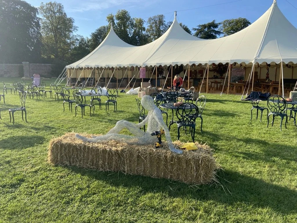 Outdoor event setup with a large white tent, black metal chairs and tables, and a hay bale with a decorative human figure sculpture made of small white lights, set in a grassy area with trees in the background.