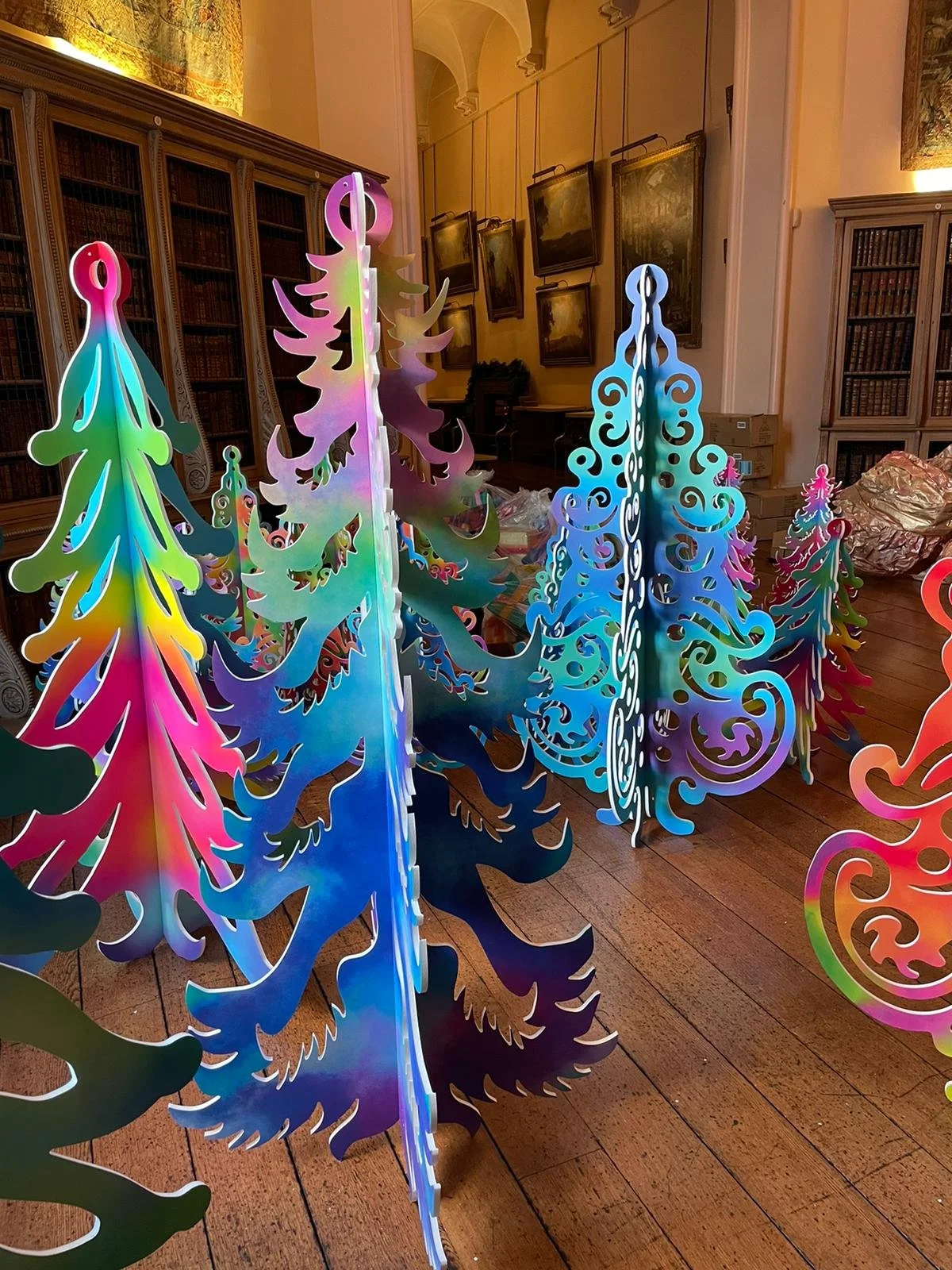 Colorful, laser-cut acrylic Christmas tree decorations displayed in a room with wooden floors, bookshelves, and framed paintings on the walls.