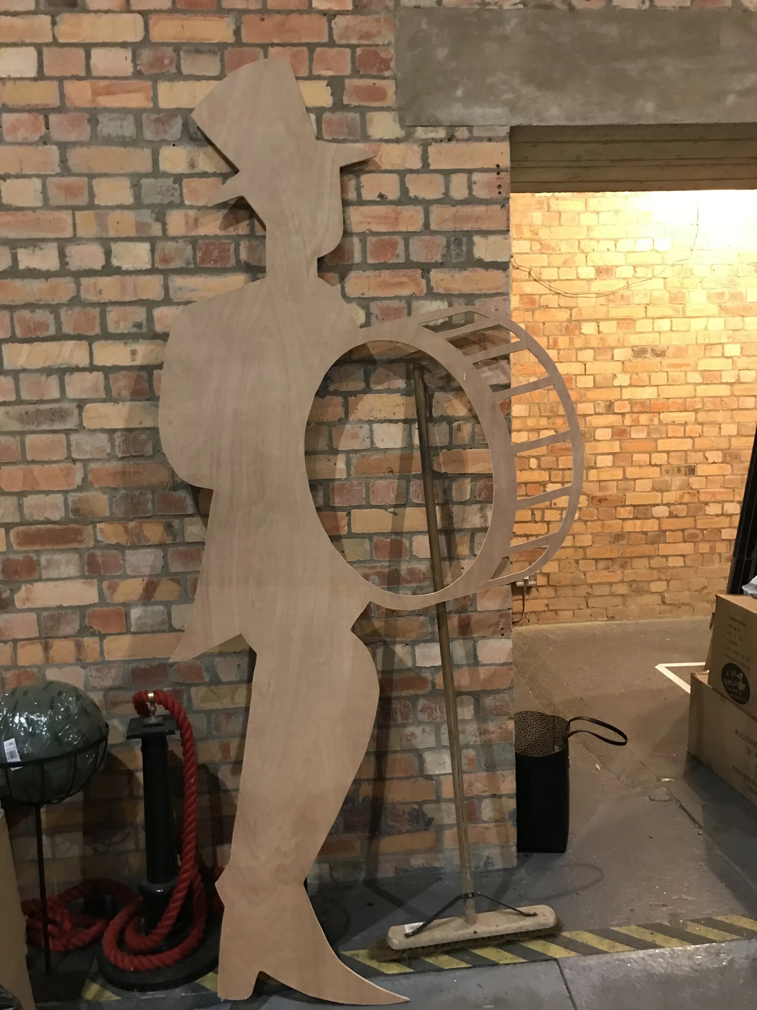 A large wooden cutout of a whimsical animal figure, likely a squirrel or a fox, is leaning against a brick wall inside a room with a concrete floor. There is a lamp, some red rope, and various objects around it, indicating it might be in a workshop o