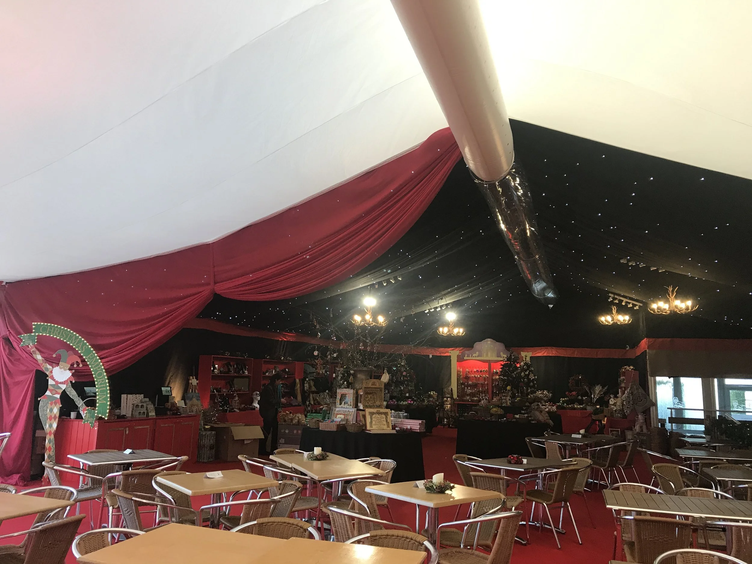 An indoor space decorated for Christmas with black drapes and star-like lights on the ceiling, red curtains, tables and chairs arranged for dining, and various Christmas display items and decorations.