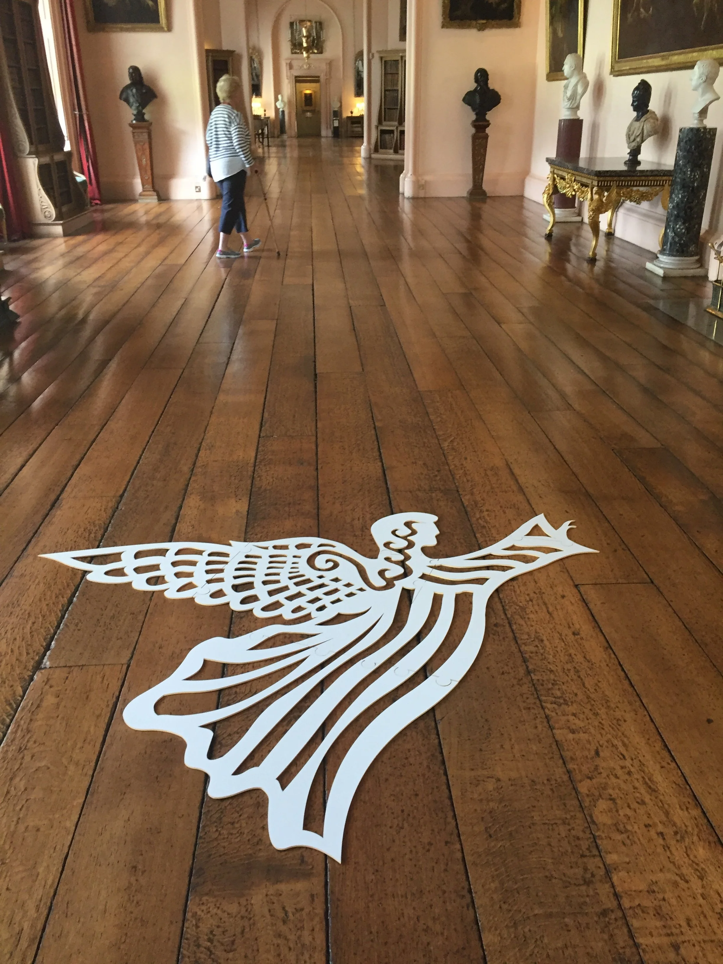 A white decorative art piece in the shape of a bird with intricate cutout patterns lies on a wooden floor in an ornate gallery or museum corridor, with a woman walking in the background.