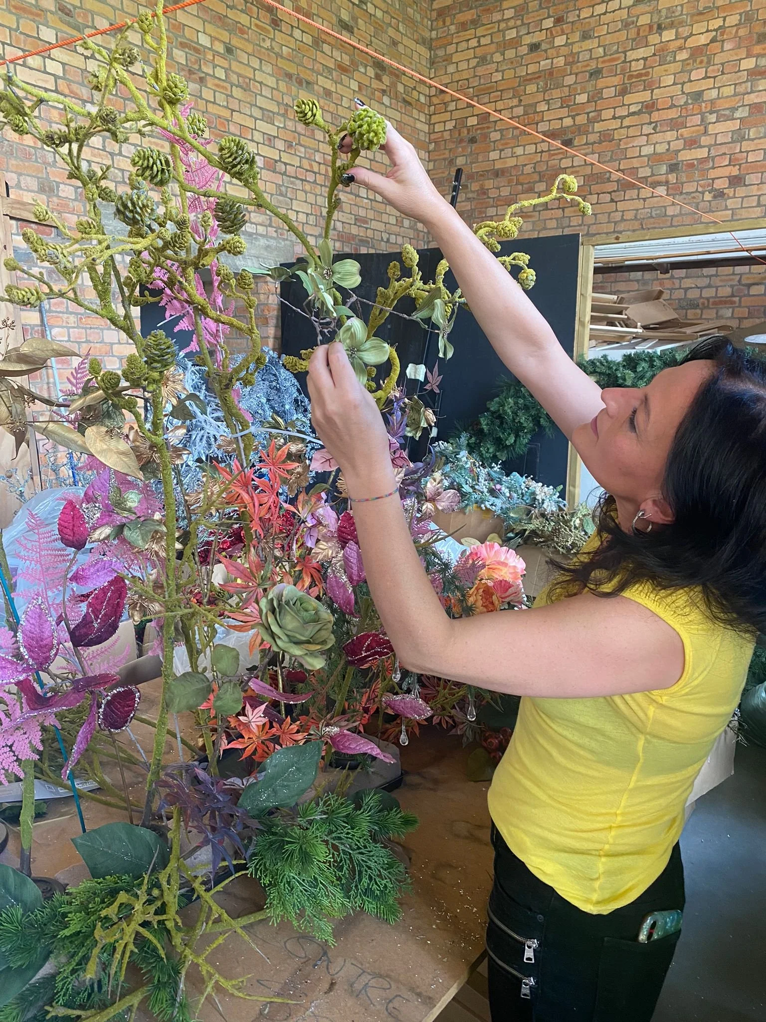 A woman in a yellow top is adjusting a decorative floral arrangement with various colorful artificial plants and flowers in an indoor setting with brick walls.