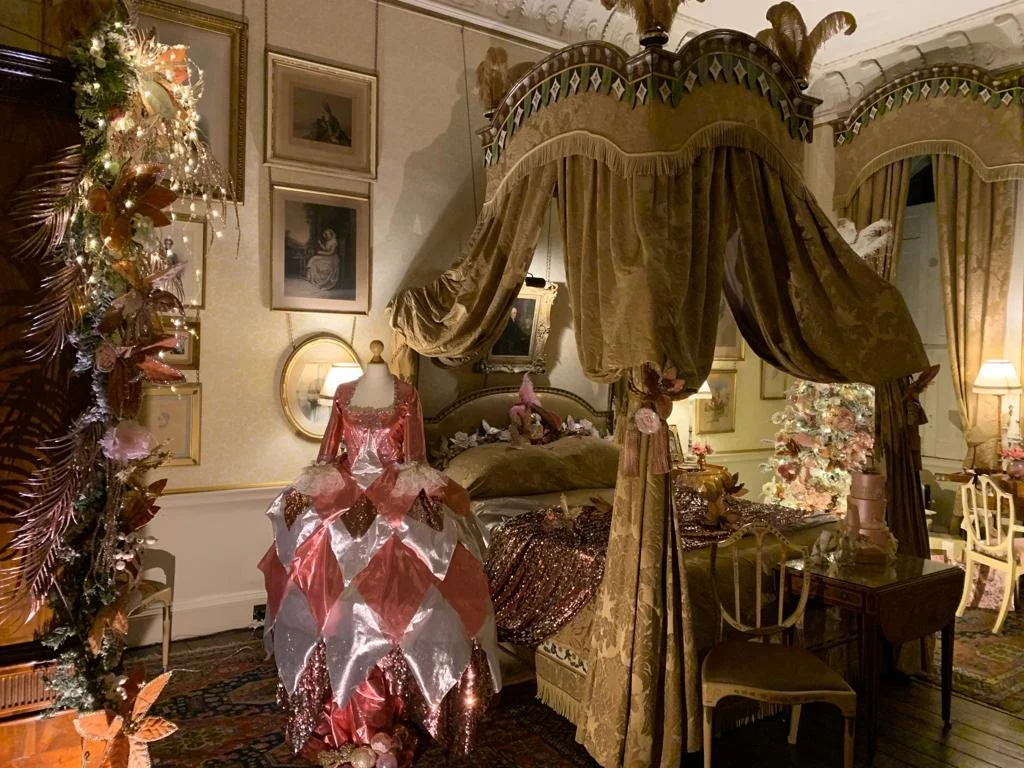 A bedroom with a four-poster bed draped with gold velvet curtains, decorated for Christmas with a Christmas tree and festive ornaments. A mannequin wearing a pink and white dress is near the bed. The walls are adorned with framed artwork, and there a