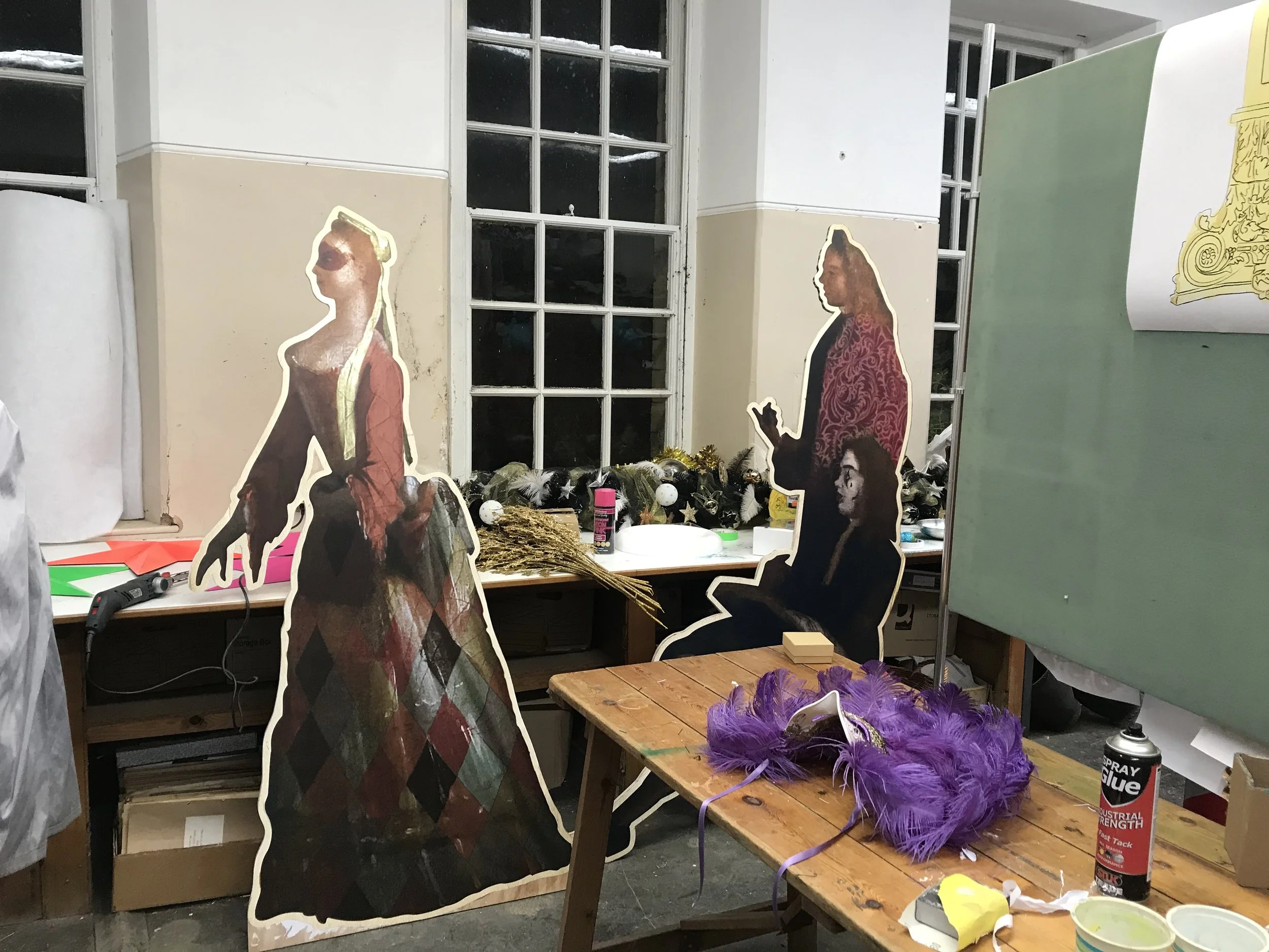 A workshop with cardboard cutouts of people, various decorations on the table, and craft supplies including feathers, glue, and spray paint.