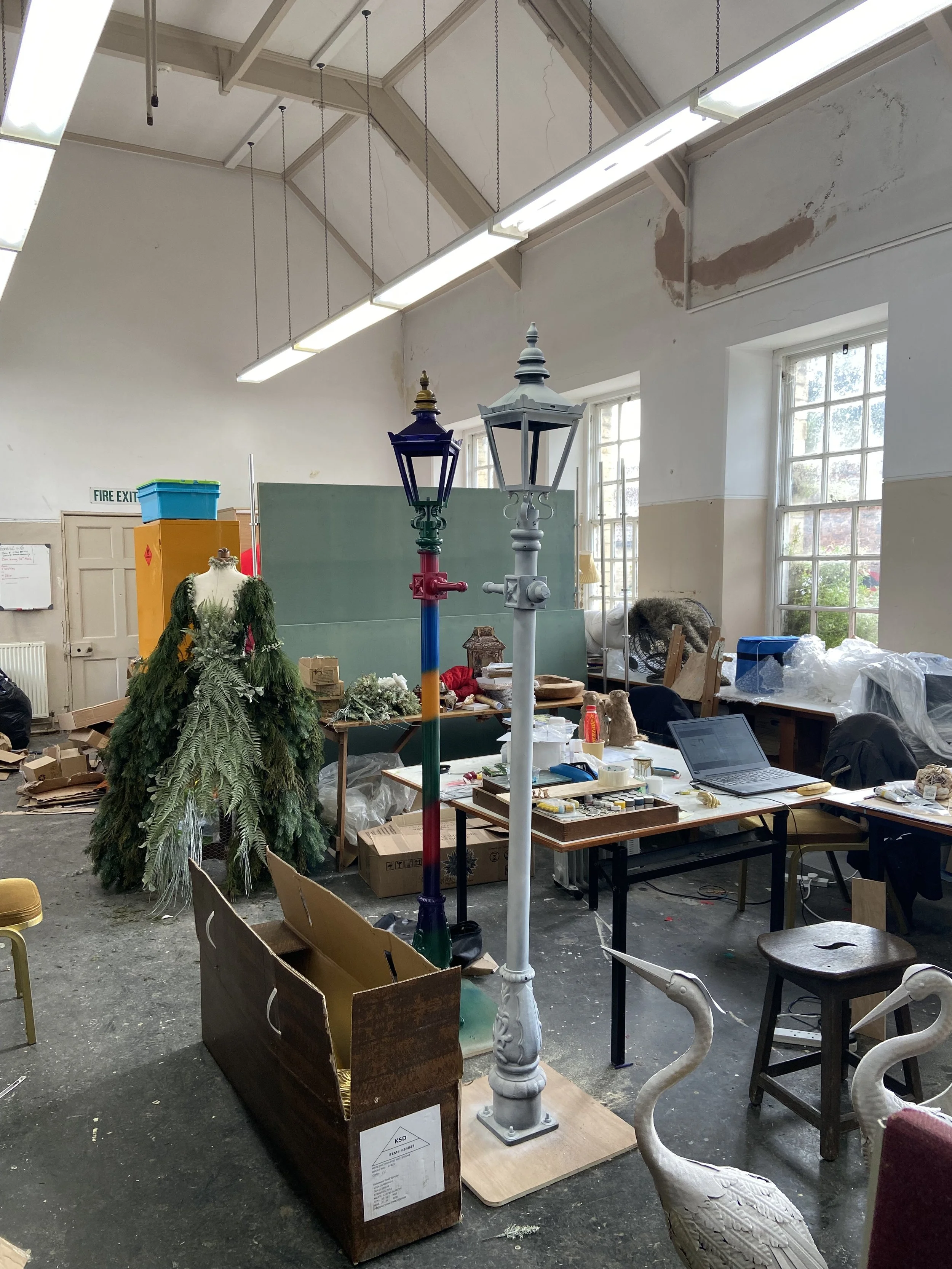 Indoor space with tables, chairs, and various decorative items including two streetlamp-style lamps, a mannequin draped with greenery, and office supplies. The room has tall windows, high ceilings, and visible wear on the walls.
