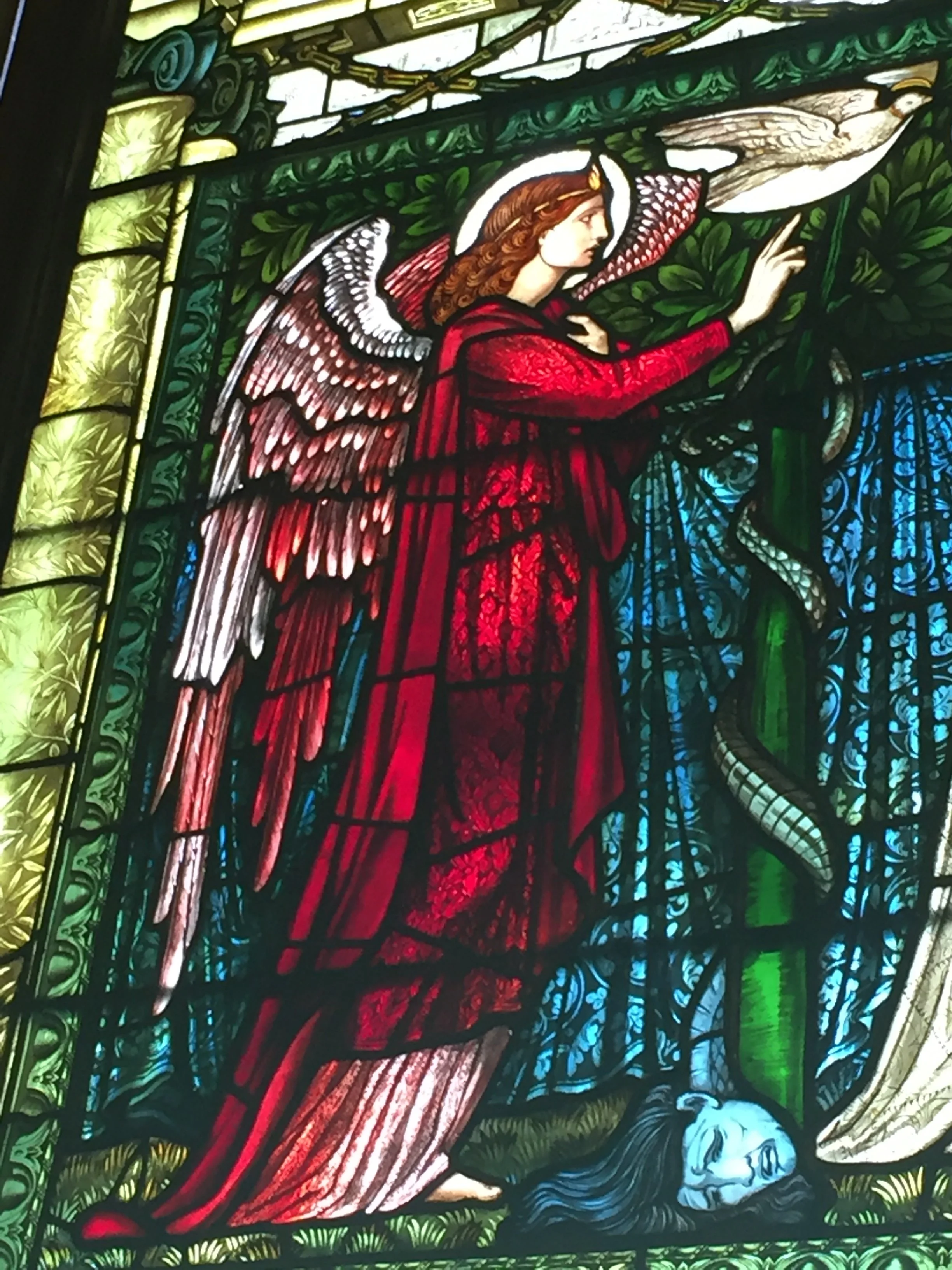 Stained glass depiction of an angel with wings and a halo, kneeling and praying over a sleeping man with long hair, in a garden setting with greenery and a flowing stream.