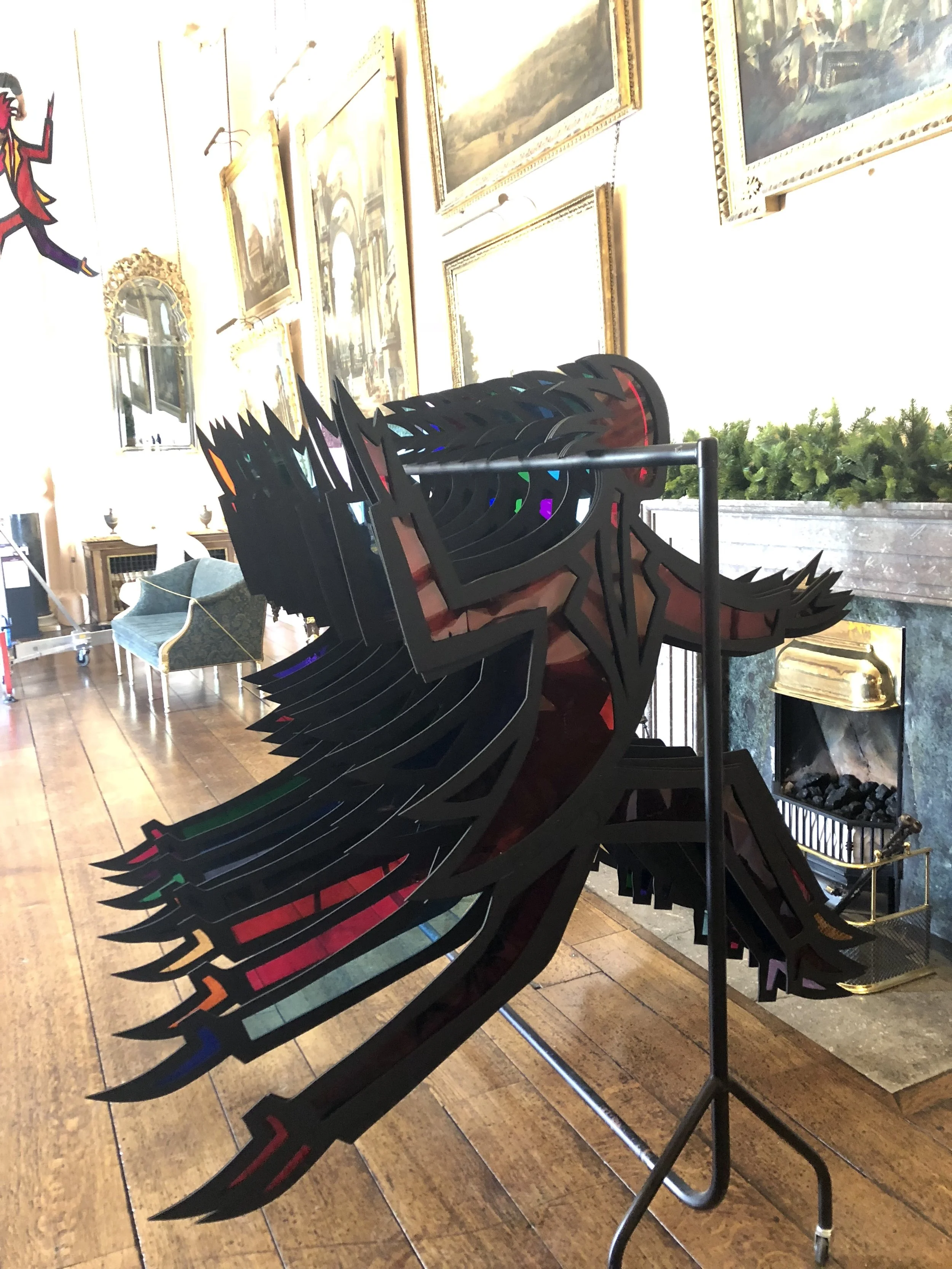 Indoor scene with framed landscape paintings on a wall and a large, black, abstract sculpture with colorful accents resembling a stylized animal or creature, placed on a metal stand on a wooden floor. There is a green cushioned armchair, a small tabl