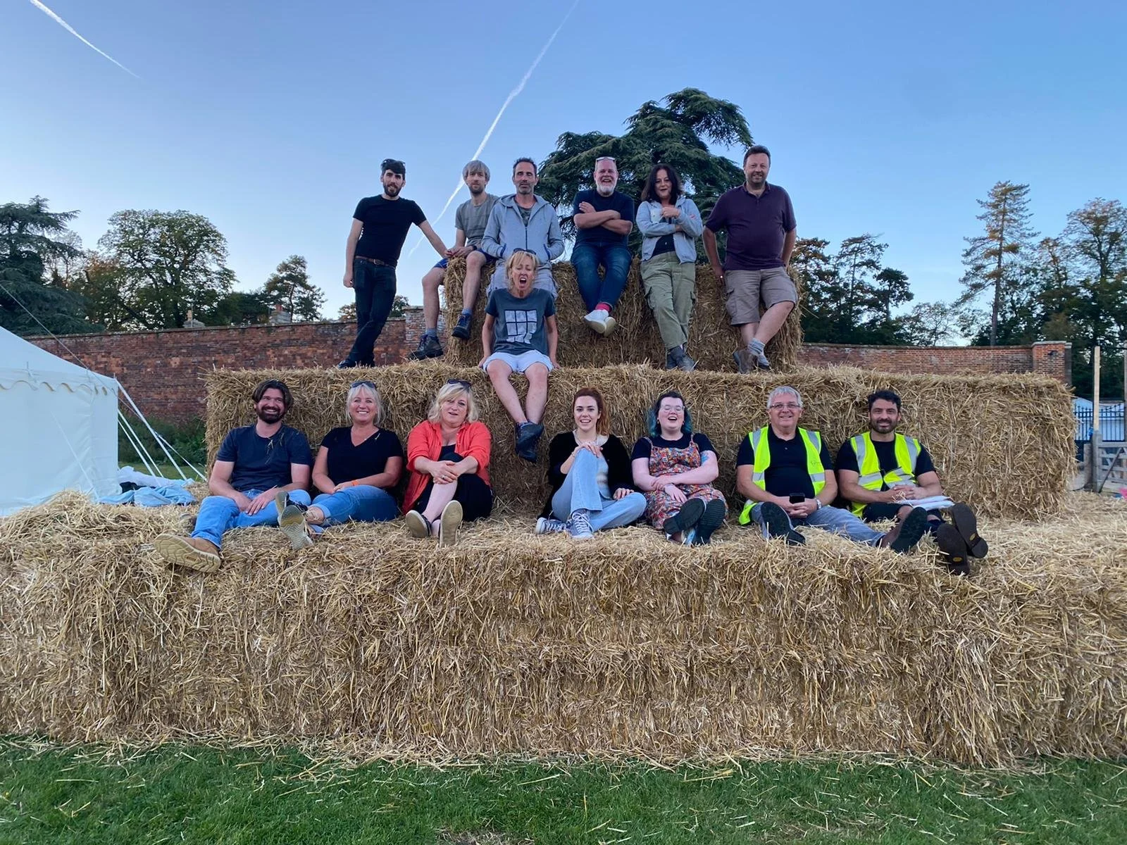 Group of people sitting and standing on large hay bales outdoors, with trees and a brick wall in the background, under a clear blue sky.