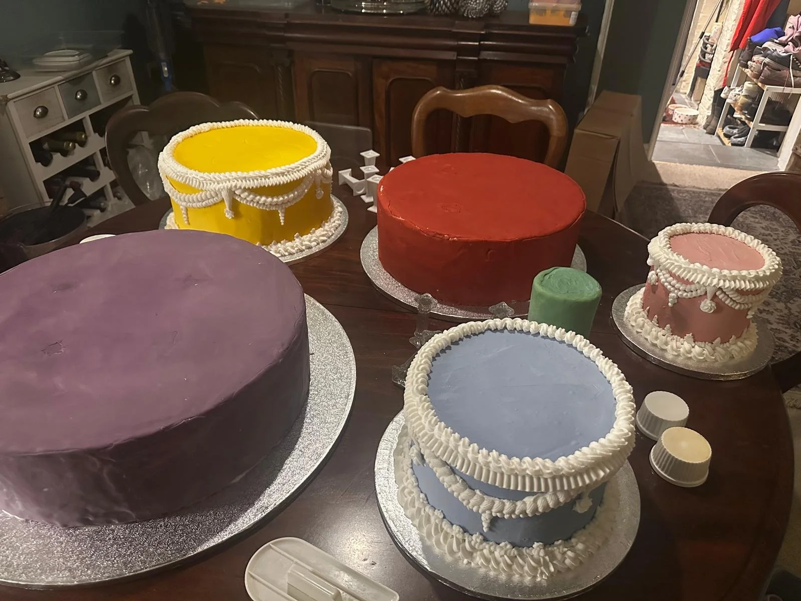 A variety of decorated cakes in different colors including purple, yellow, red, pink, and light blue, arranged on a wooden table indoors.