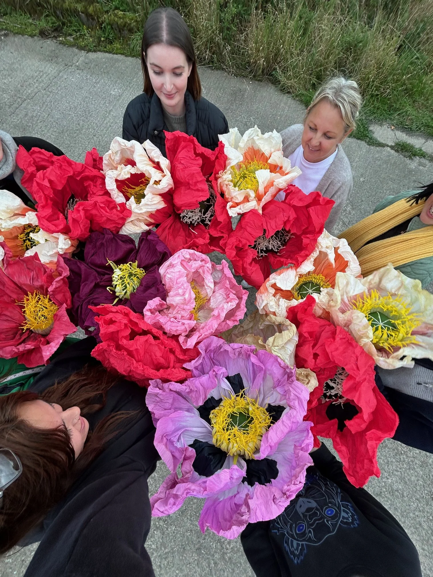 People lying on the ground surrounded by large colorful paper flowers, looking at and admiring the flowers.