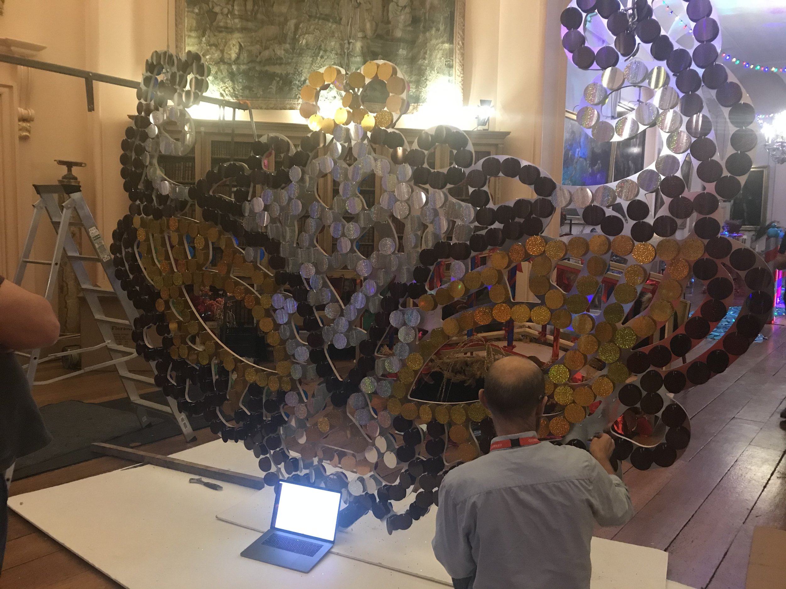 An art installation or sculpture made of numerous circular mirrors arranged in patterns and shapes, with some mirrors displaying reflective surfaces. A laptop positioned on a table displays an image or text. Several people are observing or working on