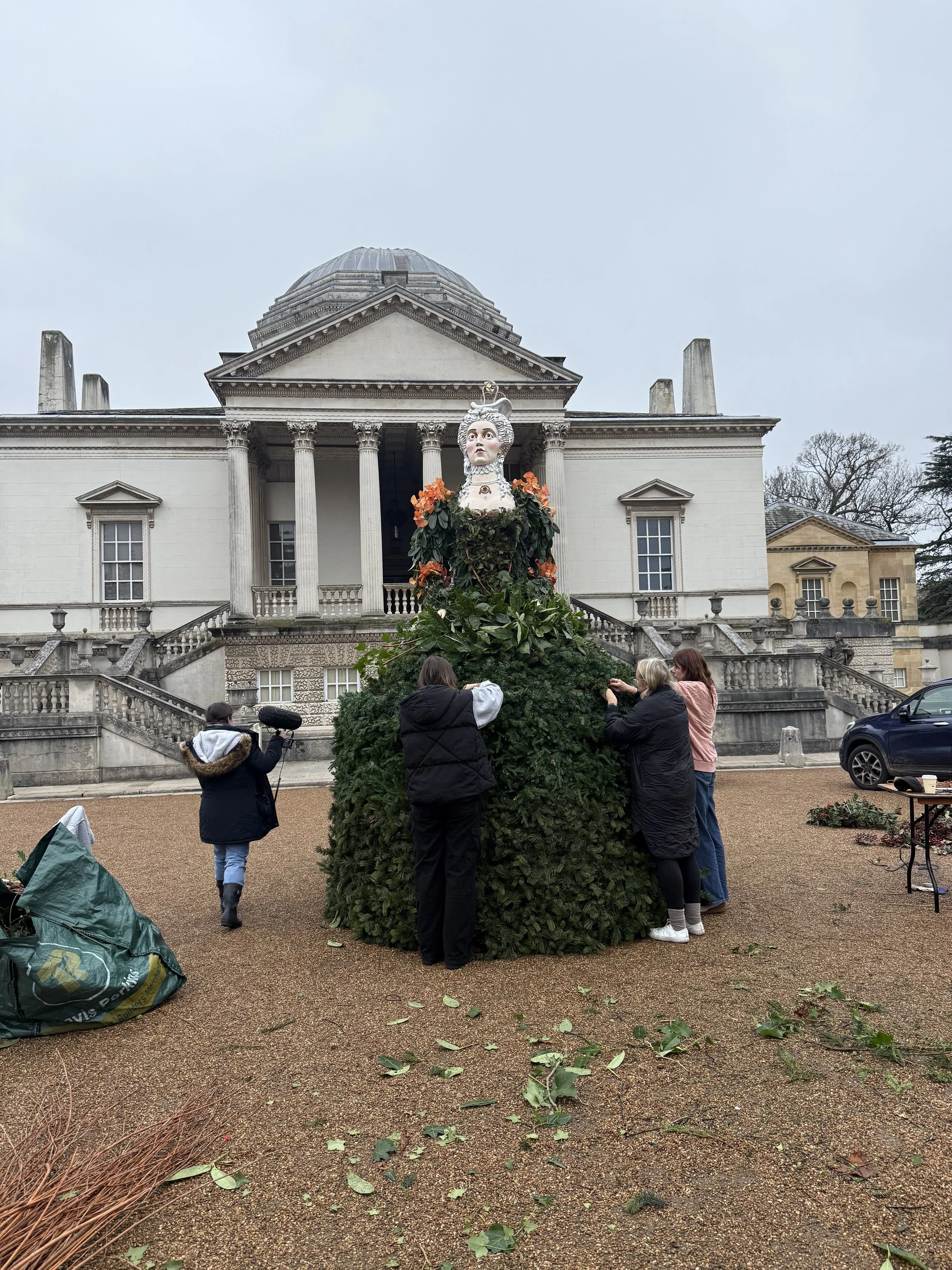 A group of people is decorating a large Christmas tree in front of a neoclassical building with columns. The tree features a large head sculpture of a woman with intricate hair and crown decorations, surrounded by artificial leaves and flowers. Green