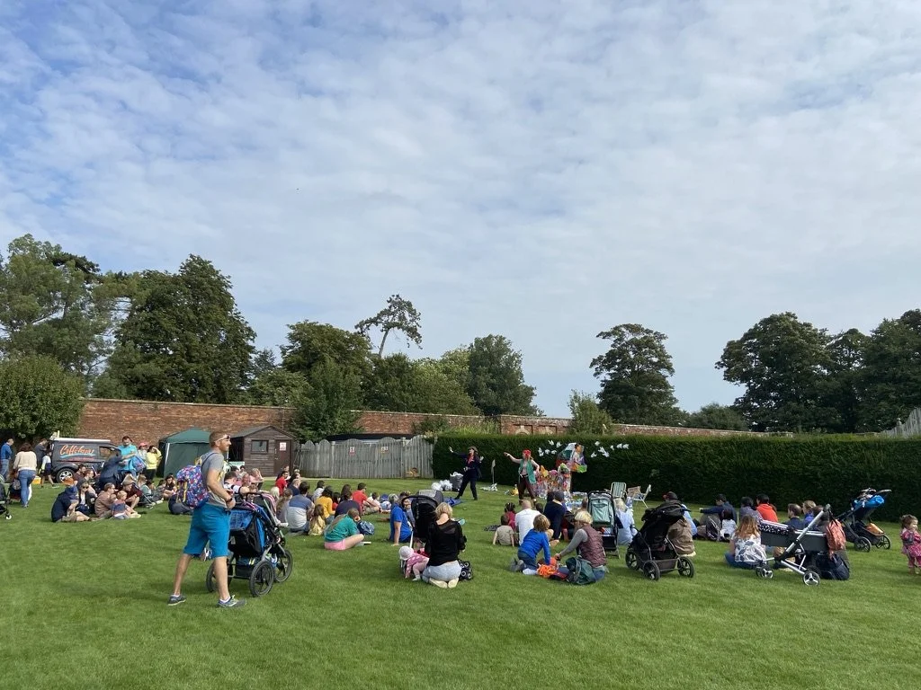 People attending an outdoor event on a grassy field, with some sitting on the grass and others pushing strollers. A performance is taking place with performers in colorful costumes and some decorations. Trees and a wall are in the background, under a