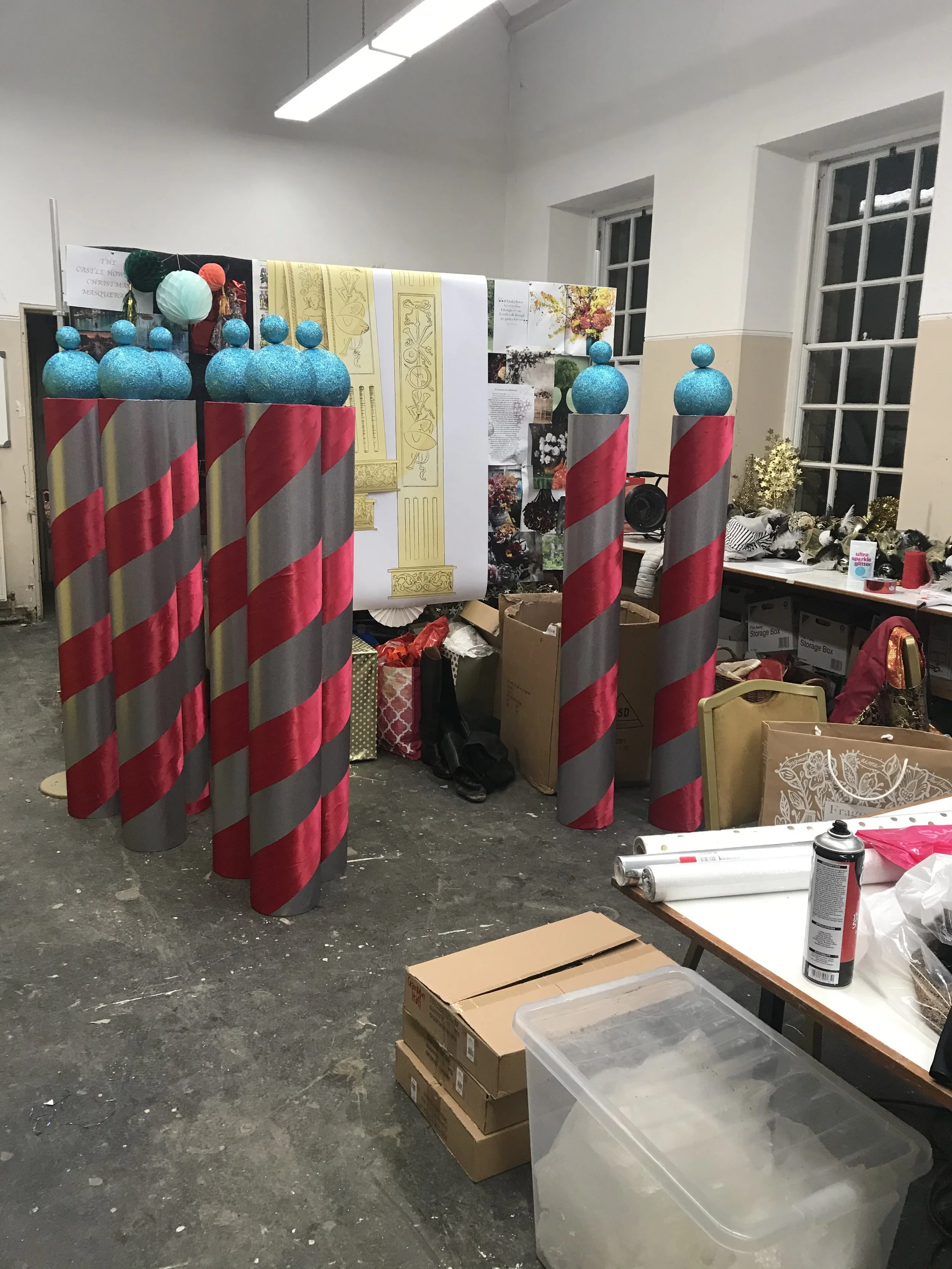 Decorations for a Christmas event, including large blue glittery baubles on top of pink and gray striped poles, hanging lanterns, and various holiday-themed items in a craft or workshop space.