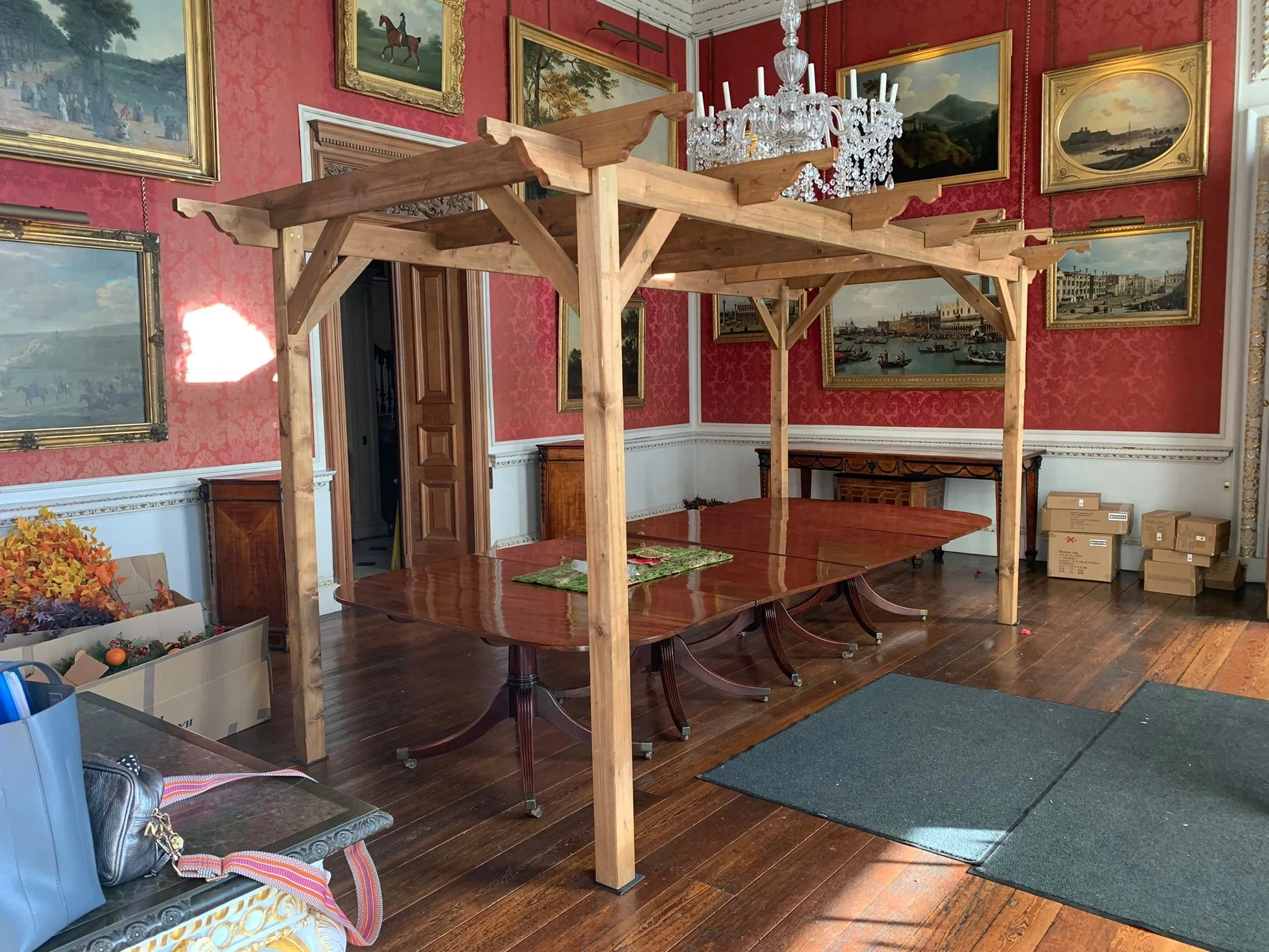 A large wooden table with a scaffold-like structure in the center of a decorated room with red patterned wallpaper, several framed landscape paintings, a chandelier, and cardboard boxes.