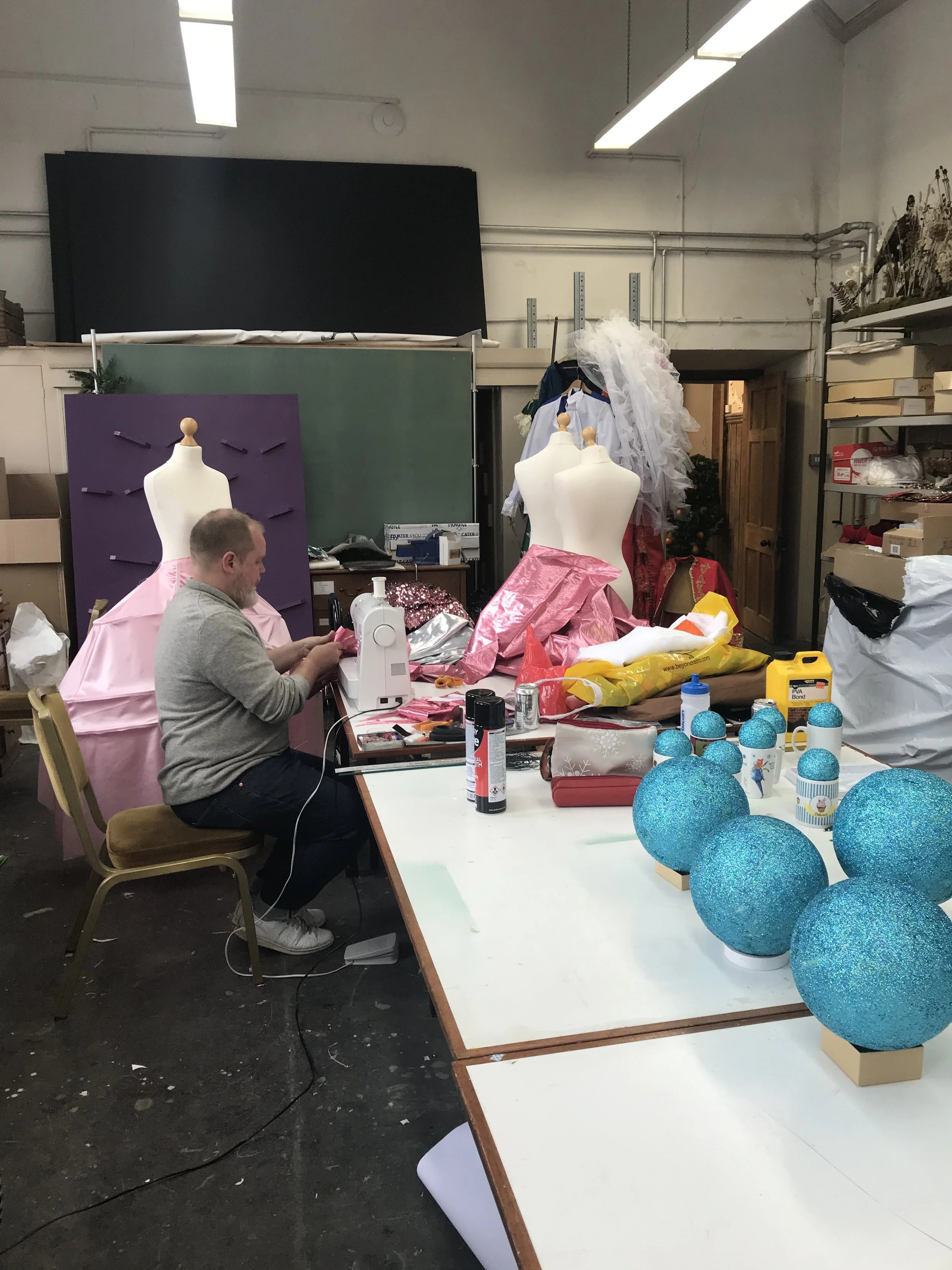 A person sewing pink fabric in a workspace filled with Christmas decorations and craft supplies, including large blue glittery ornaments on the table.