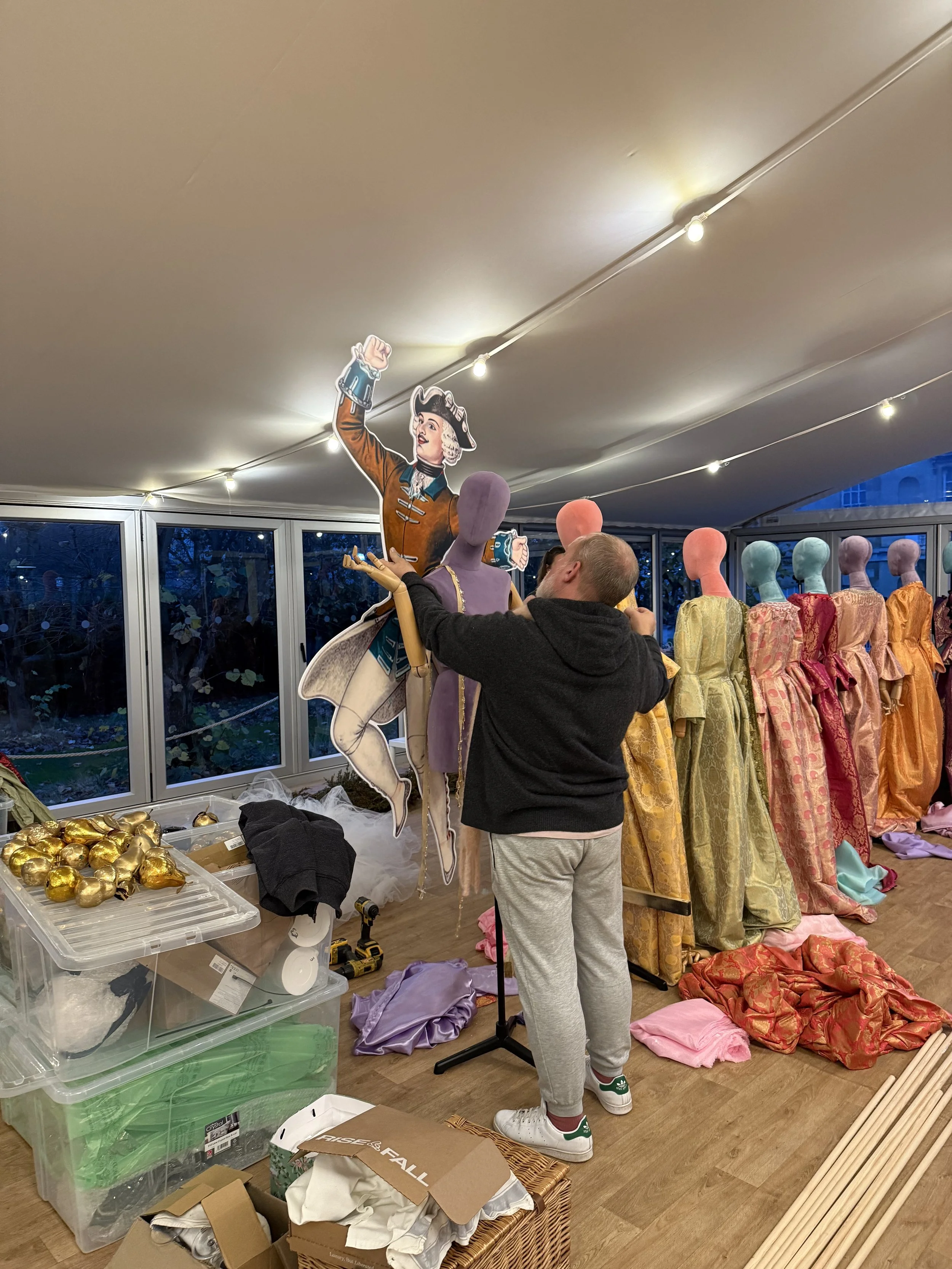A man arranging mannequins dressed in colorful clothing at an indoor event or sale. The setting includes display tables, shelves of gold-wrapped items, and scattered fabrics on the floor.