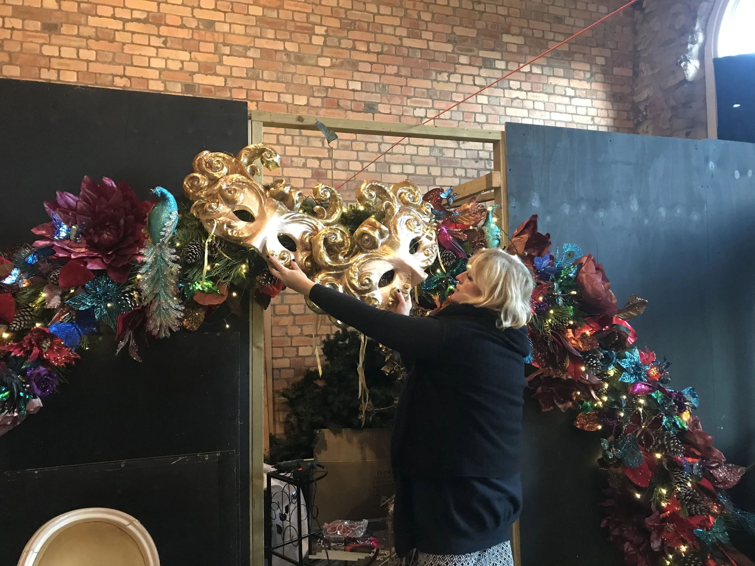 A woman decorating a large Christmas wreath adorned with shiny ornaments, poinsettias, and lights, in front of a brick wall with a window on the right.