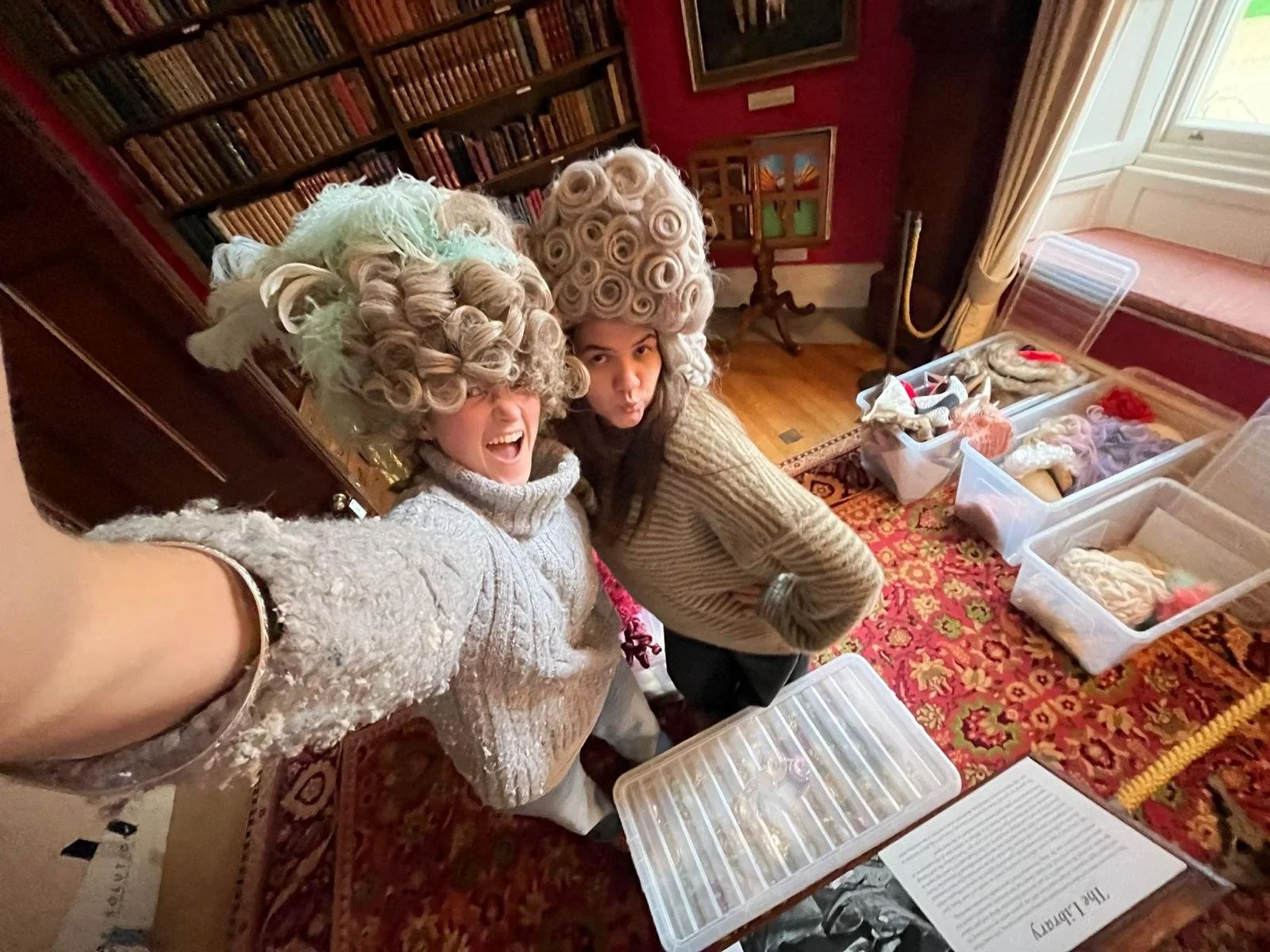 Two women wearing elaborate, curly, powdered wigs take a selfie in a room filled with bins of fabric and sewing supplies. The room has a colorful rug and bookshelves.