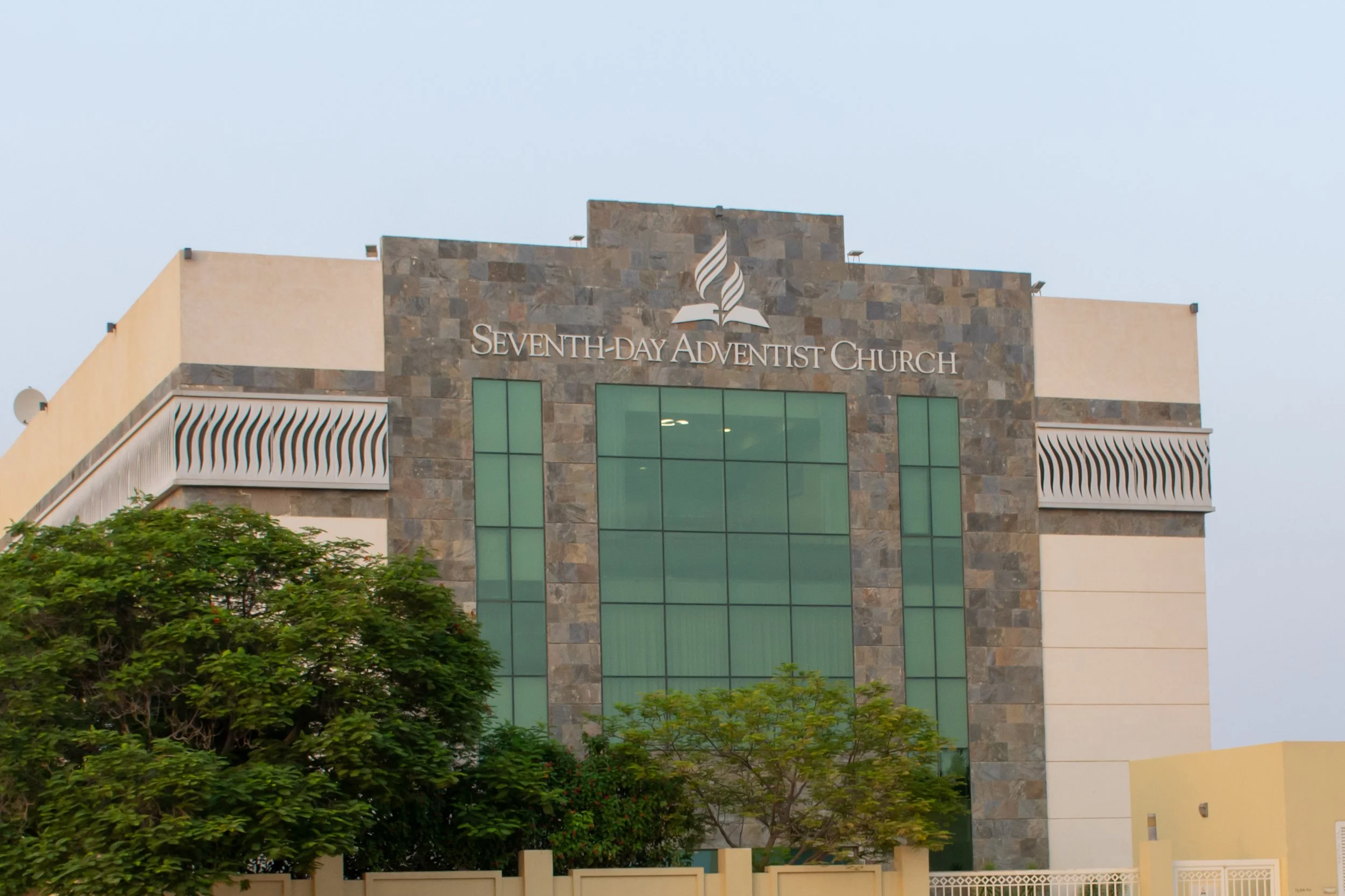 Photo of the Seventh-day Adventist Church building with stone and glass facade, trees in the foreground, and a clear sky in the background.