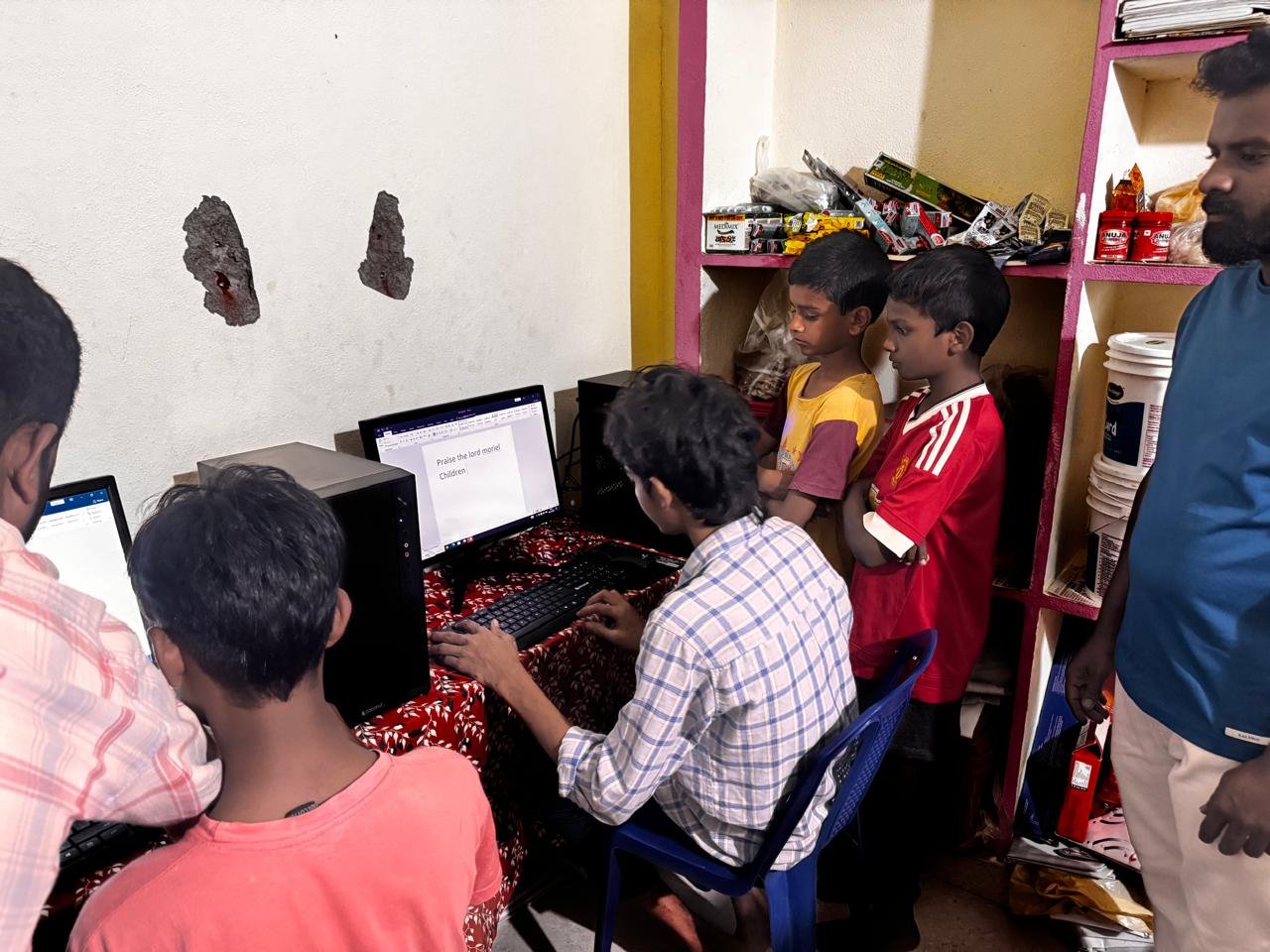A group of boys and a man gather around computers in a small room, with snacks on shelves in the background. One boy is typing on a keyboard while others watch.