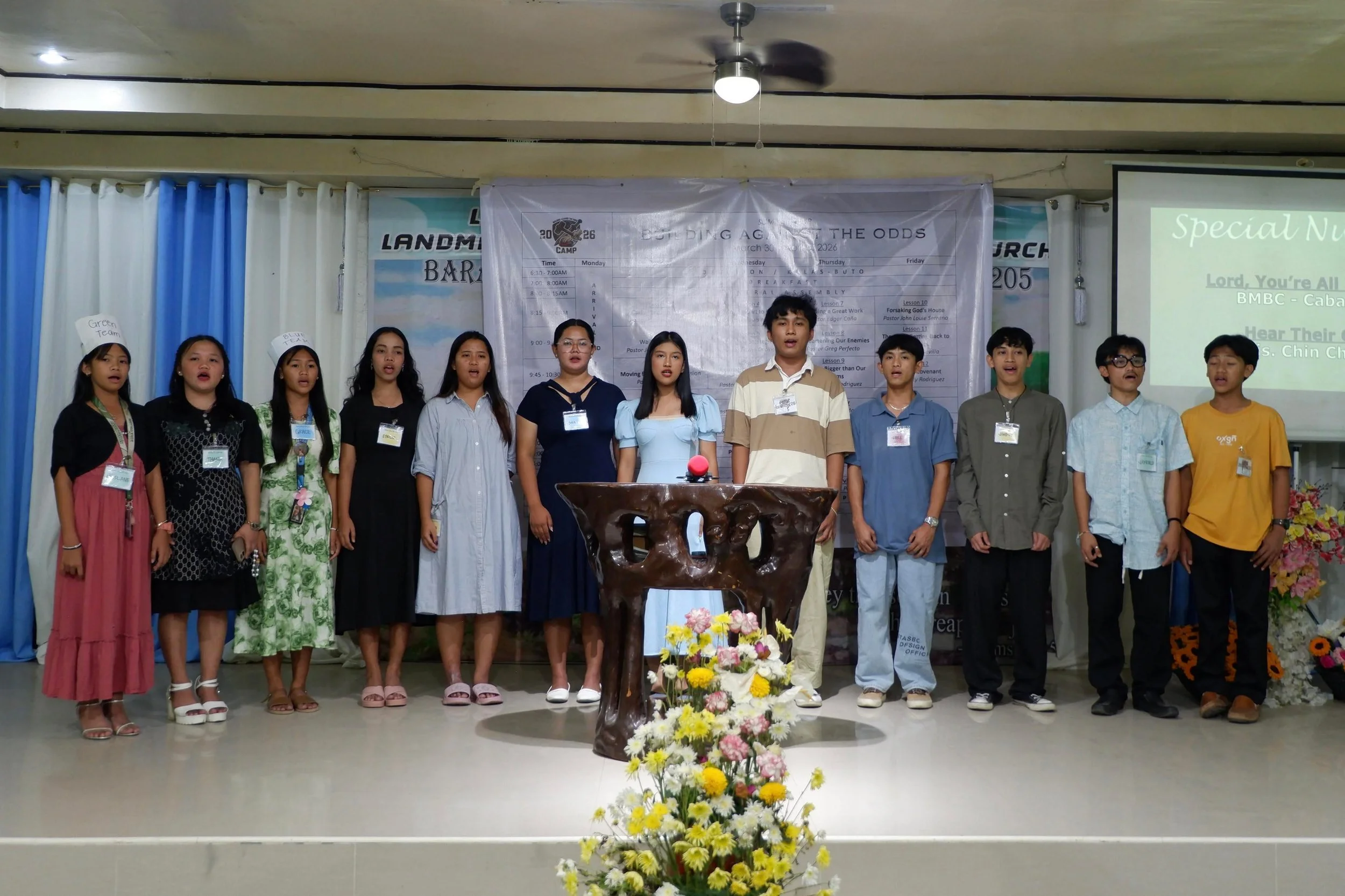Group of young people standing on stage in front of a large white banner with text and a logo, some wearing name tags, with a flower arrangement in front of the stage and a projection screen to the right.