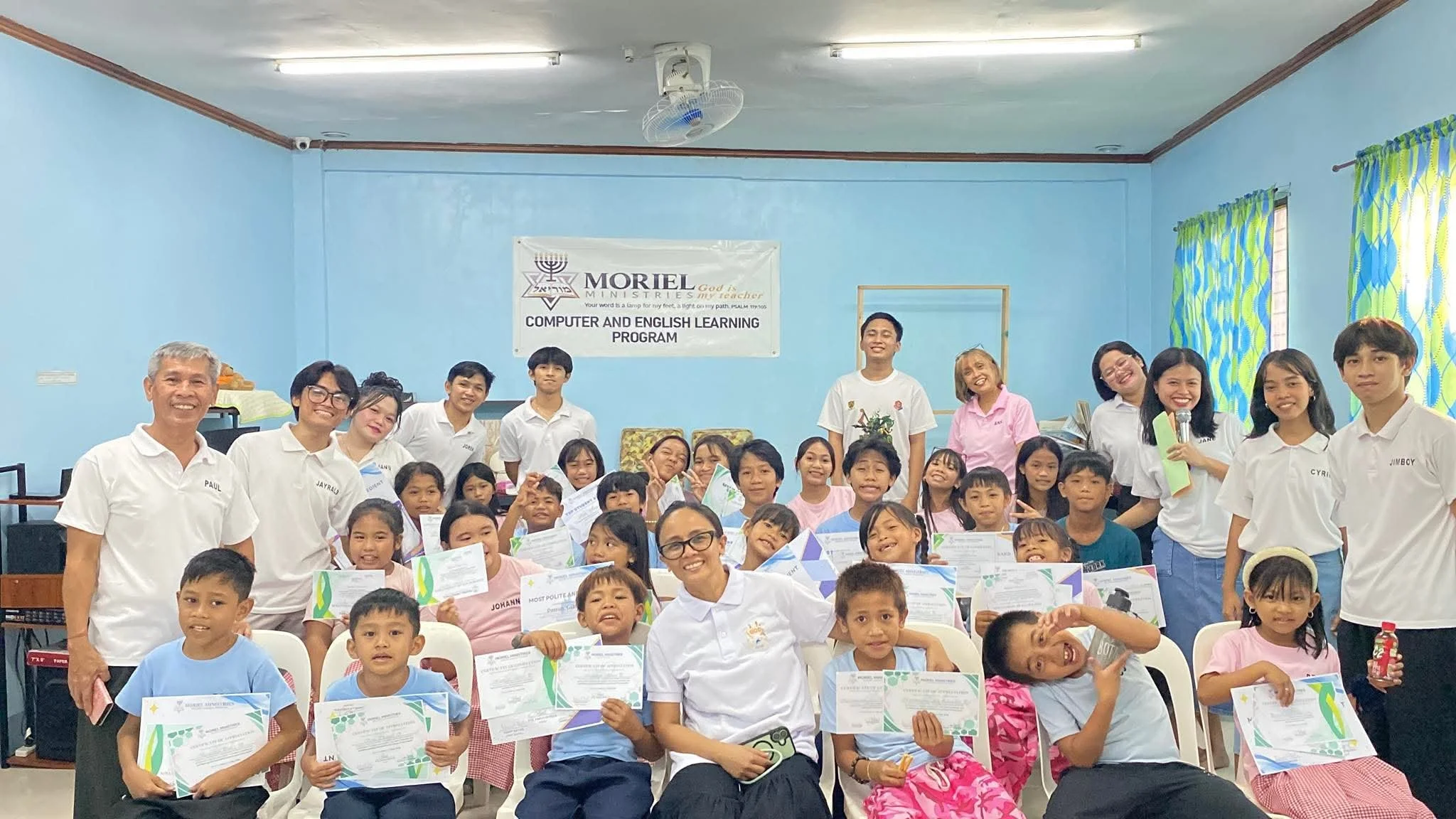 Group photo of children and adults at a learning program event, with a banner in the background reading 'MORIEL Ministries, Computer and English Learning Program' in a blue-walled room.