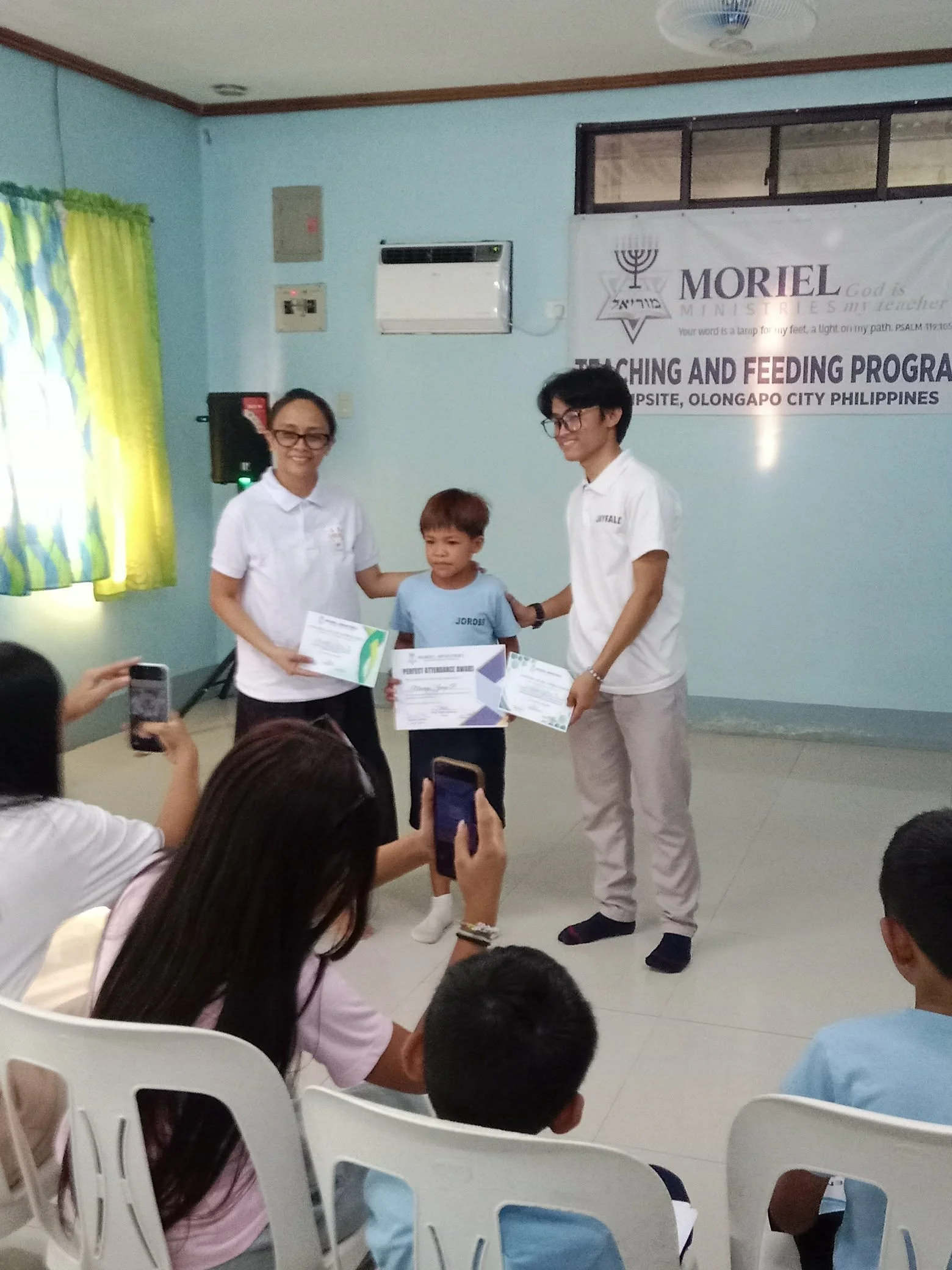 Three individuals standing in front of a light blue wall with a banner behind them. One young boy receiving certificates from two adults, one woman and one man, while other children sit in white chairs and take pictures.