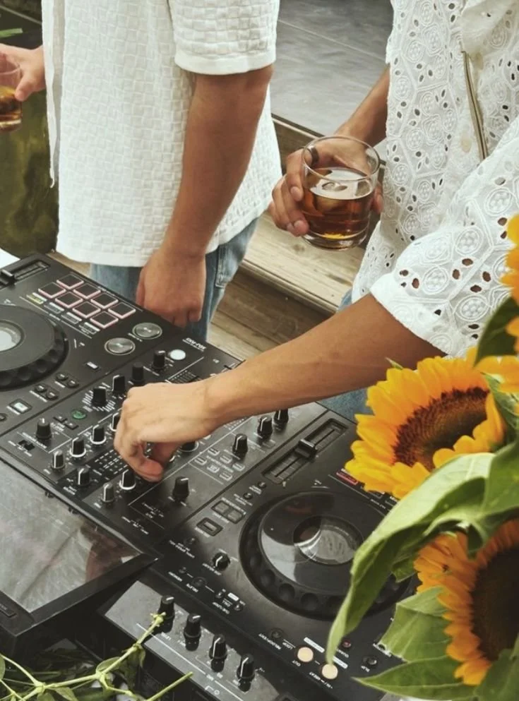 People gathered around a DJ equipment with one person adjusting knobs, another holding a glass of dark beverage, and sunflower decorations visible.