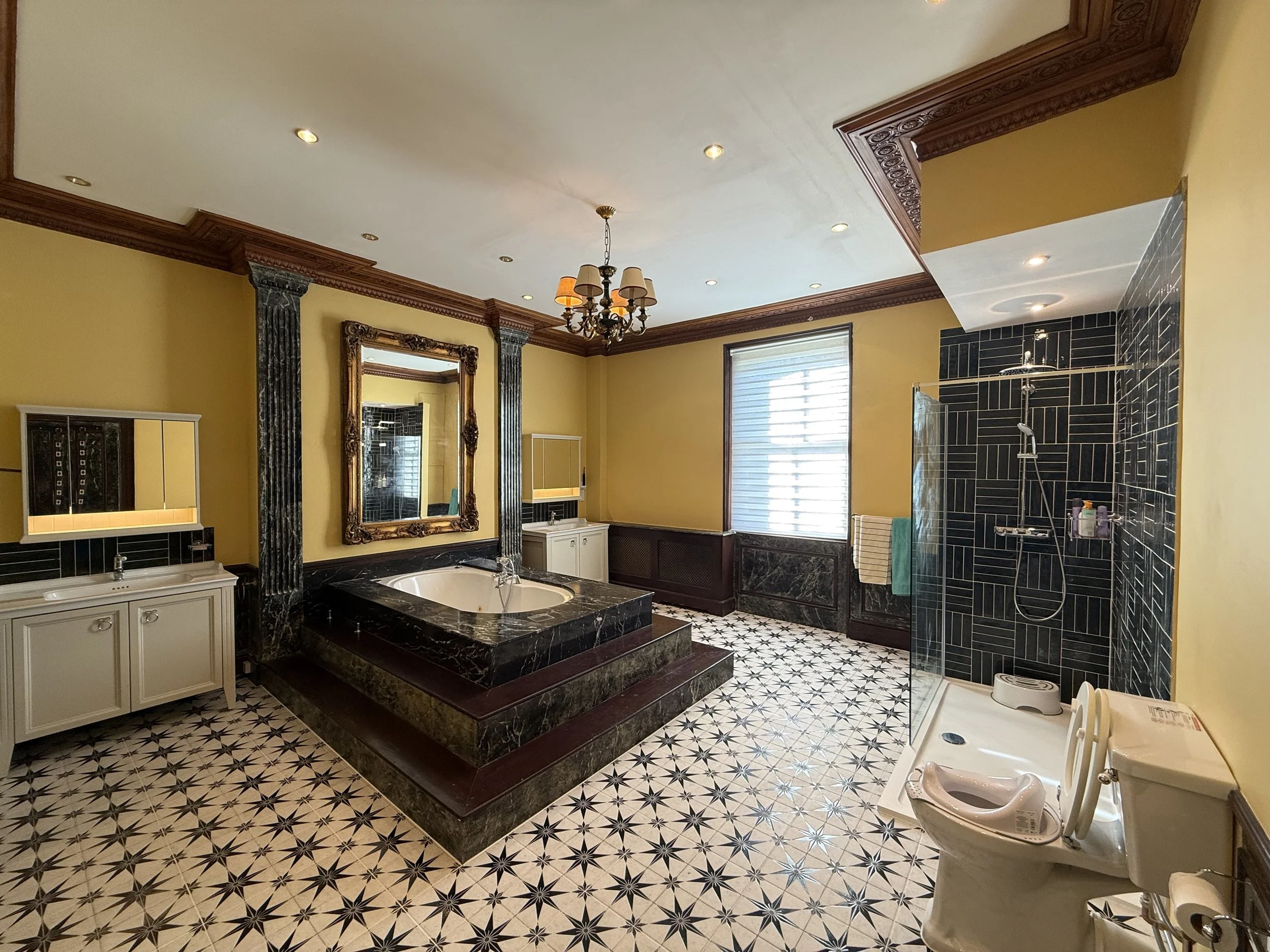 Luxury bathroom with a black marble bathtub, gold-framed mirror, and black tiled shower area, yellow walls, and patterned floor tiles.