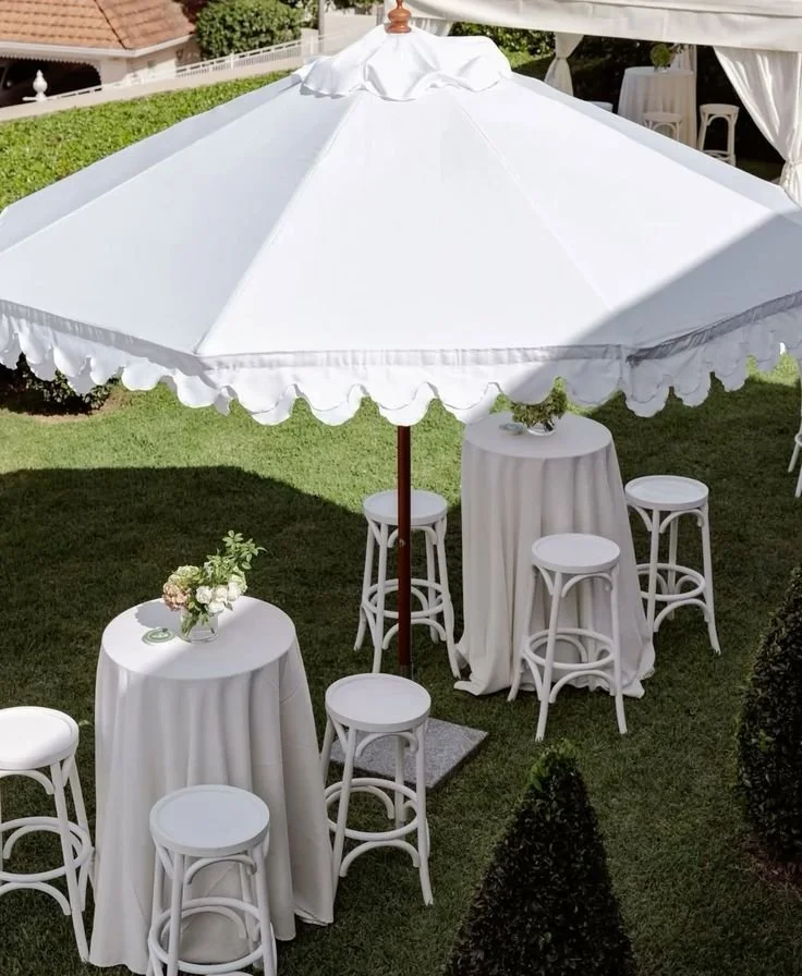 Outdoor wedding or event setup with a large white patio umbrella, tall round tables with white tablecloths, white stools, and a floral centerpiece.