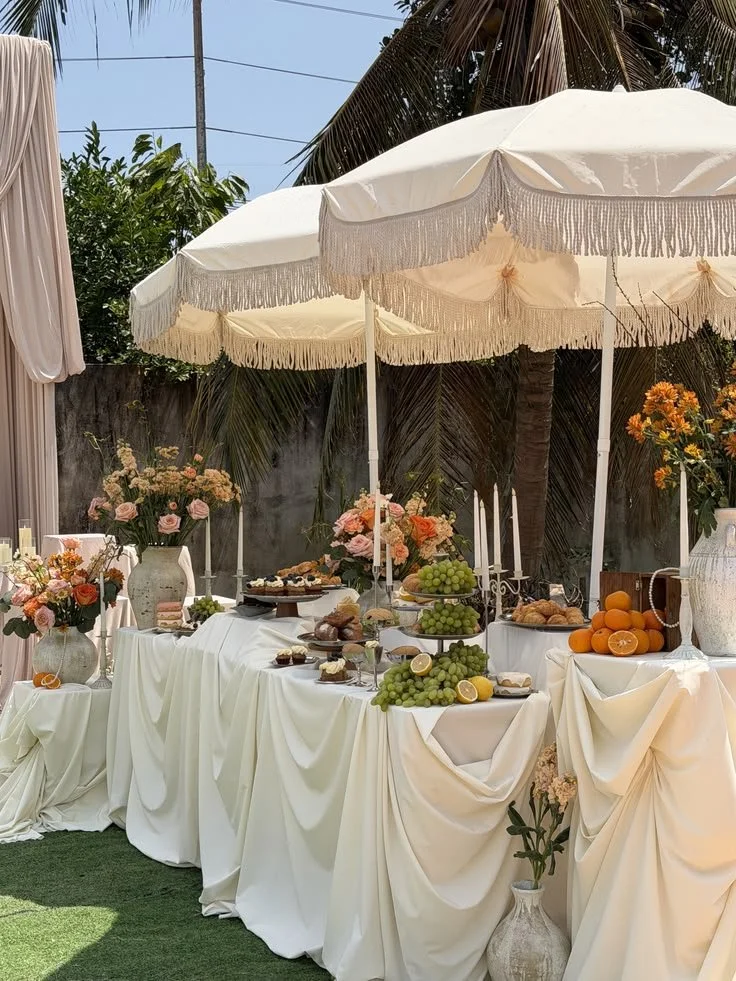 A beautifully decorated outdoor table with white draped tablecloths, large white umbrellas with fringes, and floral arrangements. The table features various desserts, fruits including grapes and oranges, and decorative candles.