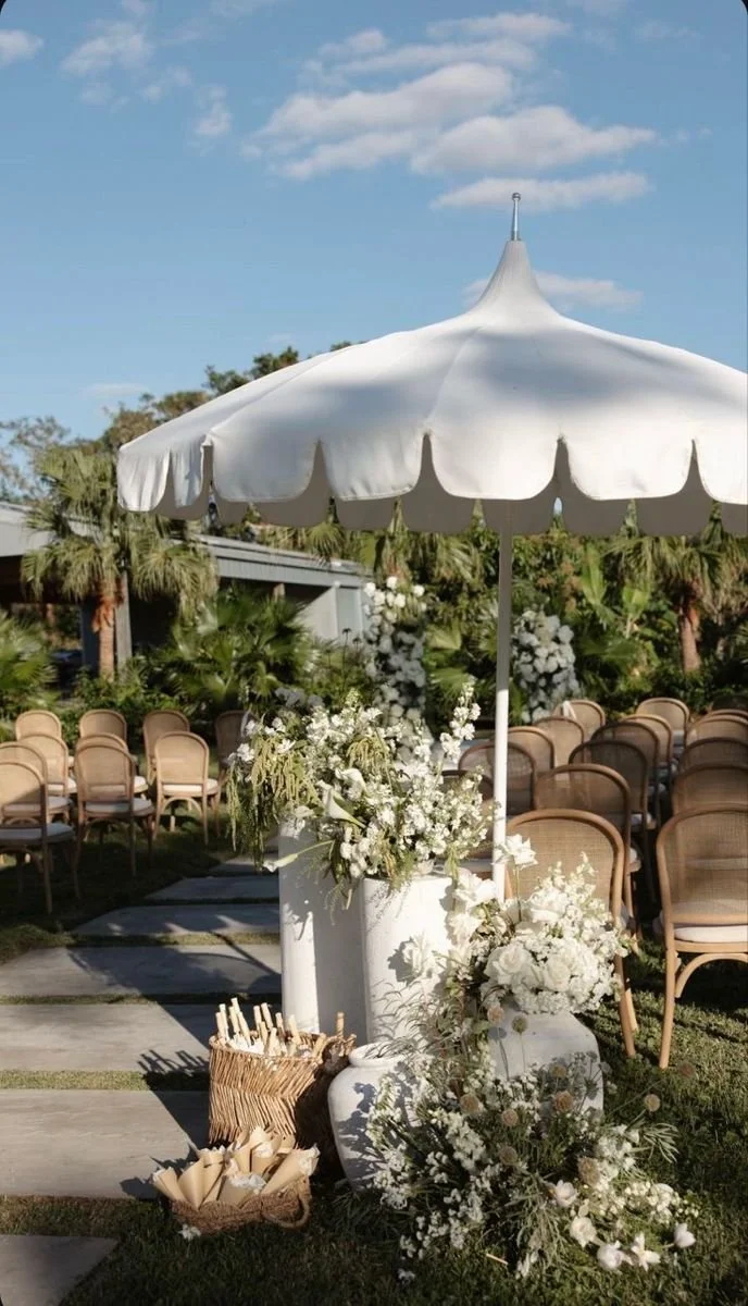 White outdoor wedding setup with a large umbrella, floral arrangements, and chairs arranged in rows.