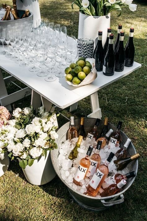 Table with wine bottles, green apples, empty wine glasses, and a potted plant with white flowers, set outdoors on grass.