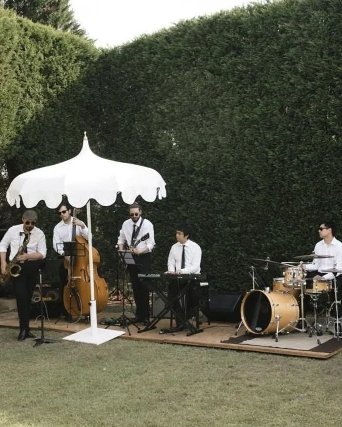 A live band performing outdoors under a white patio umbrella, with four musicians playing various instruments including saxophone, double bass, keyboard, and drums, in front of a tall hedge.