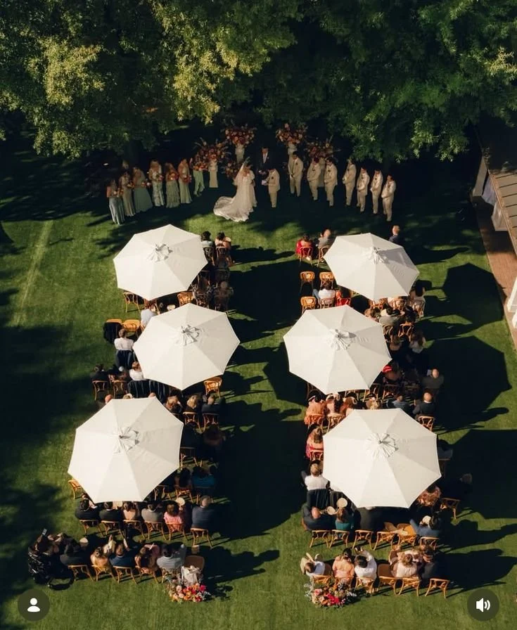View of a wedding ceremony taking place outdoors on a grassy area, with guests seated under white umbrellas and the bride and groom standing in front of a large group of people beneath trees.
