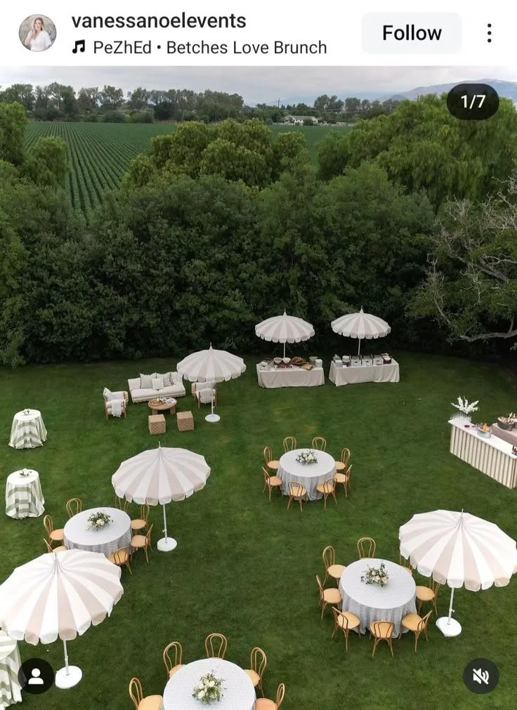 Setup for outdoor brunch with round tables covered with white tablecloths, wooden chairs, large white umbrellas, a lounge area with a couch and ottomans, and buffet tables with food and decor, all on a green lawn surrounded by trees and open fields.