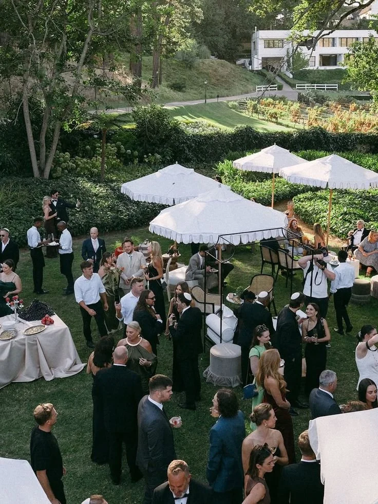 People attending an outdoor wedding reception or party in a lush garden with white umbrellas and tables, surrounded by greenery and trees.