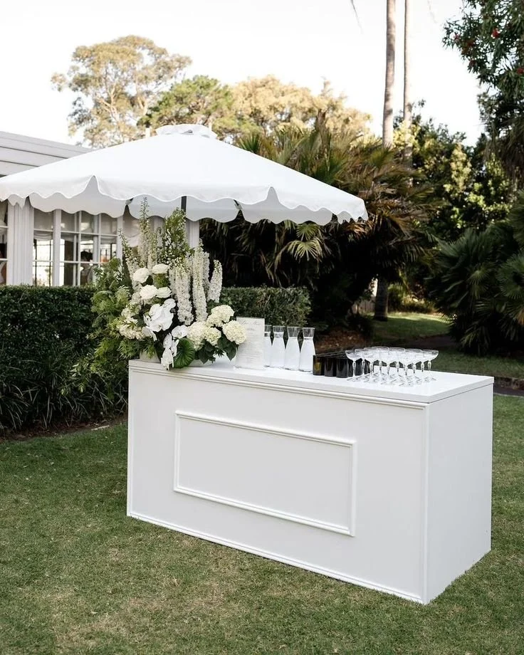 A white outdoor bar with a floral arrangement, under a white umbrella, set up on a lawn with trees and bushes in the background.