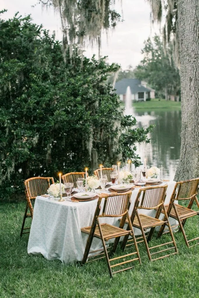 beautifully arranged wooden table & chairs with candles set next to a lake with a fountain