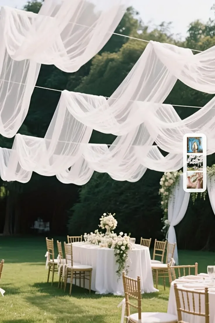 Outdoor event setup with a round table decorated with white flowers and surrounded by gold chairs, under flowing white fabric draped from strings in a garden setting.
